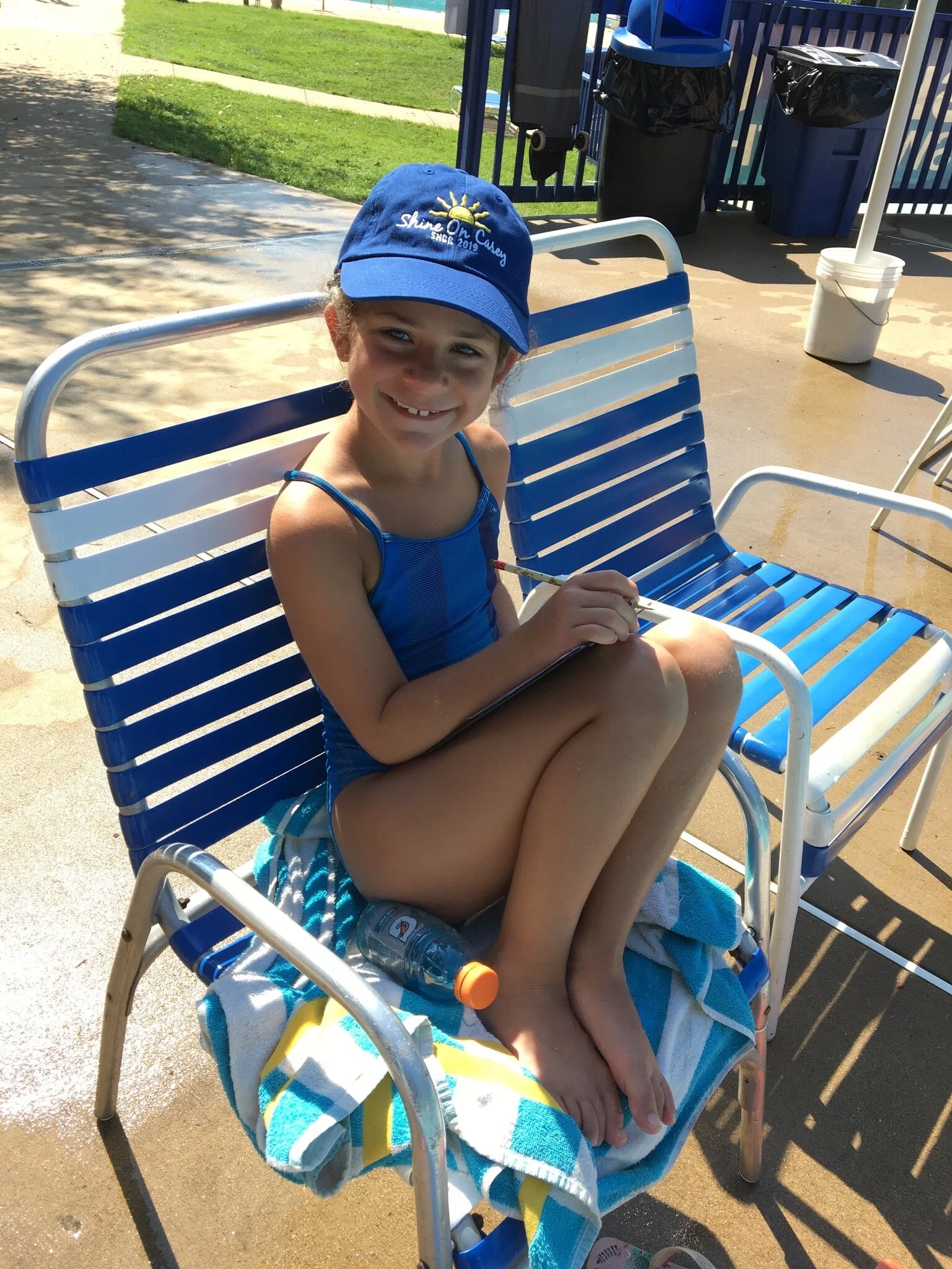 Young girl sitting on a lounge chair by the pool, holding a clipboard and pencil, wearing a blue swimsuit, a blue cap, and smiling at the camera.