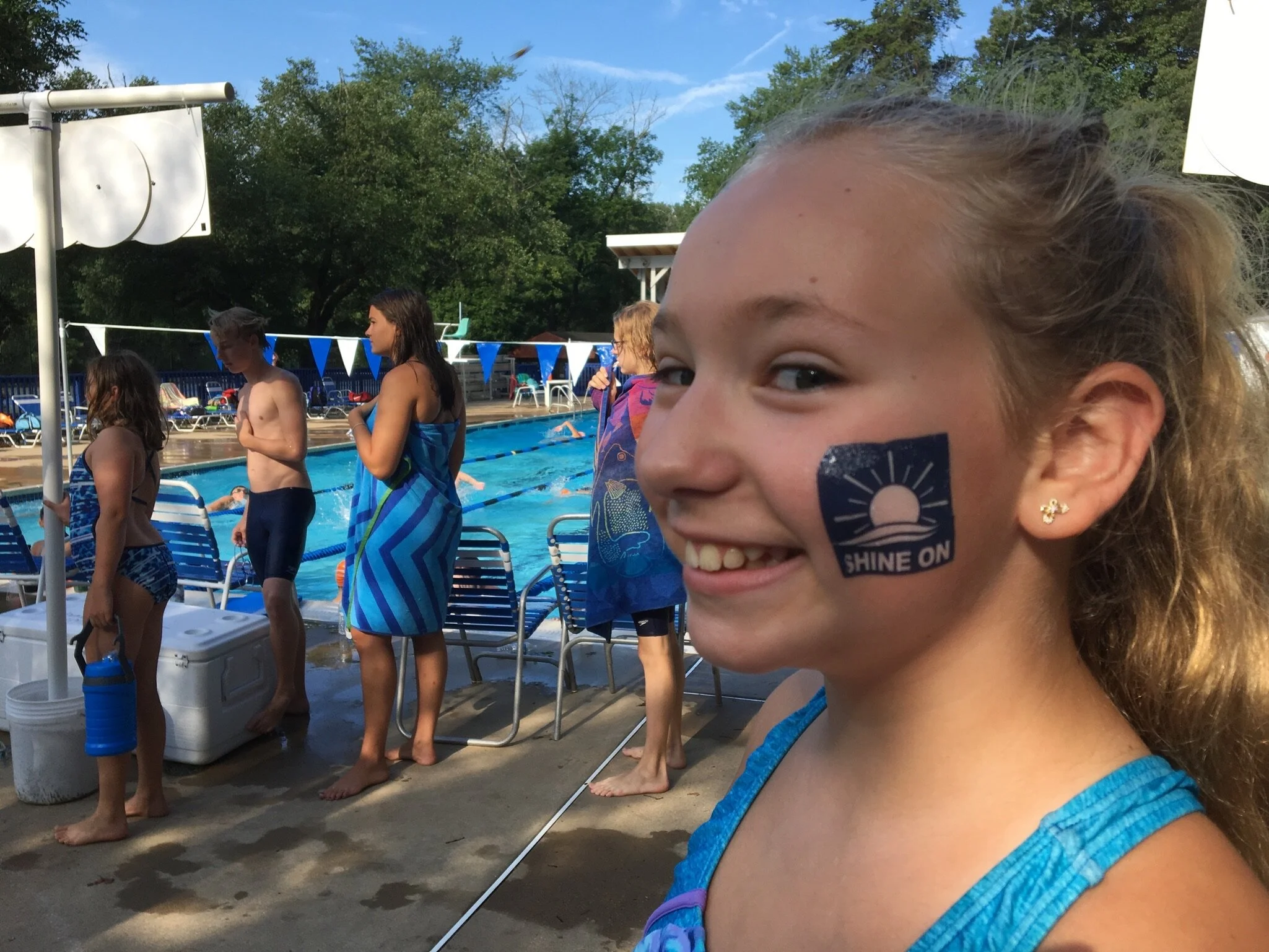 A young girl with a temporary tattoo on her cheek that says 'Shine On' and a picture of a sunset or sunrise. She is smiling at a swimming pool with other children and adults in swimsuits, under blue flags, on a sunny day with trees in the background.