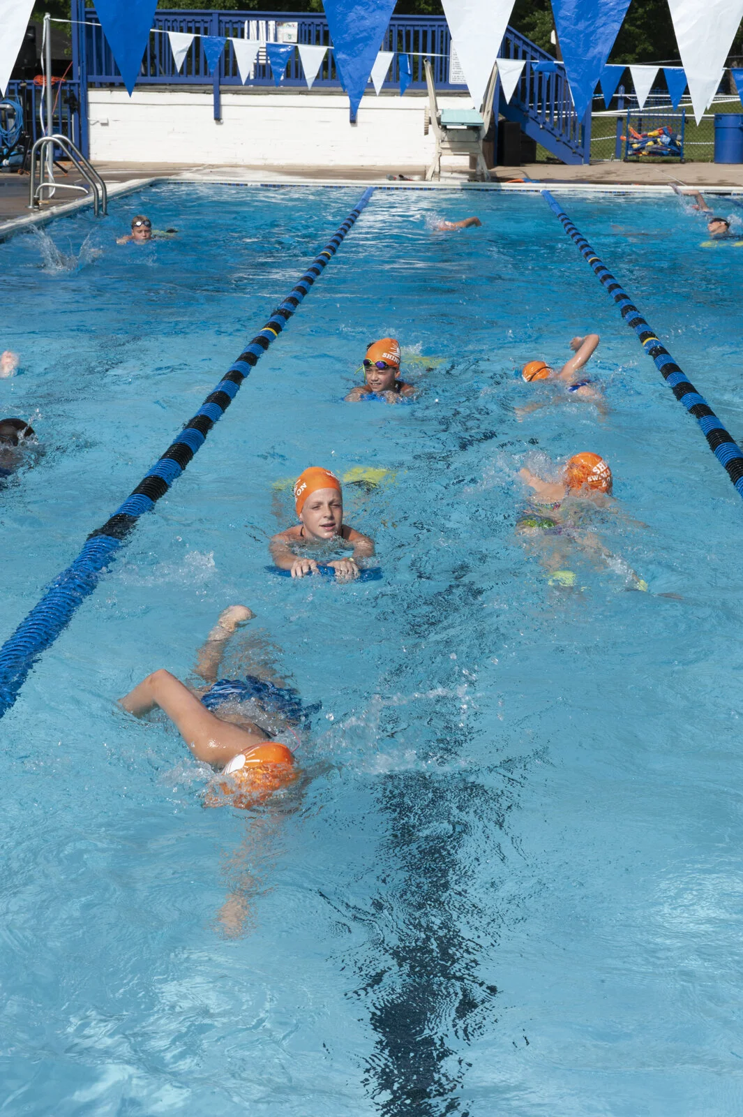 Children swimming in a lap pool with blue and white lane dividers, some wearing orange swim caps, at an outdoor swimming facility.
