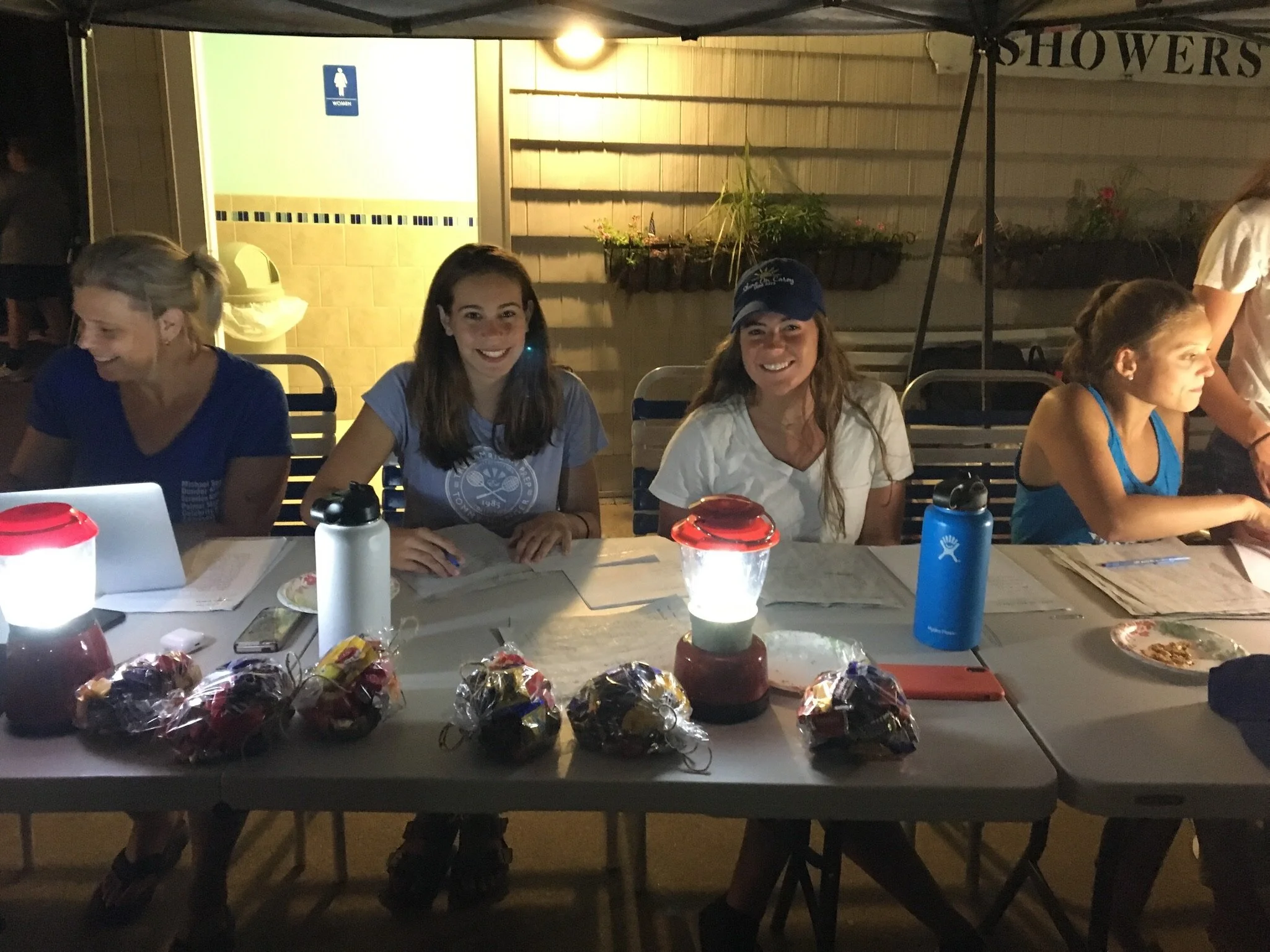 Four women sitting at a table outdoors at night, with bags of snacks, water bottles, and paper on the table, smiling and enjoying time together.