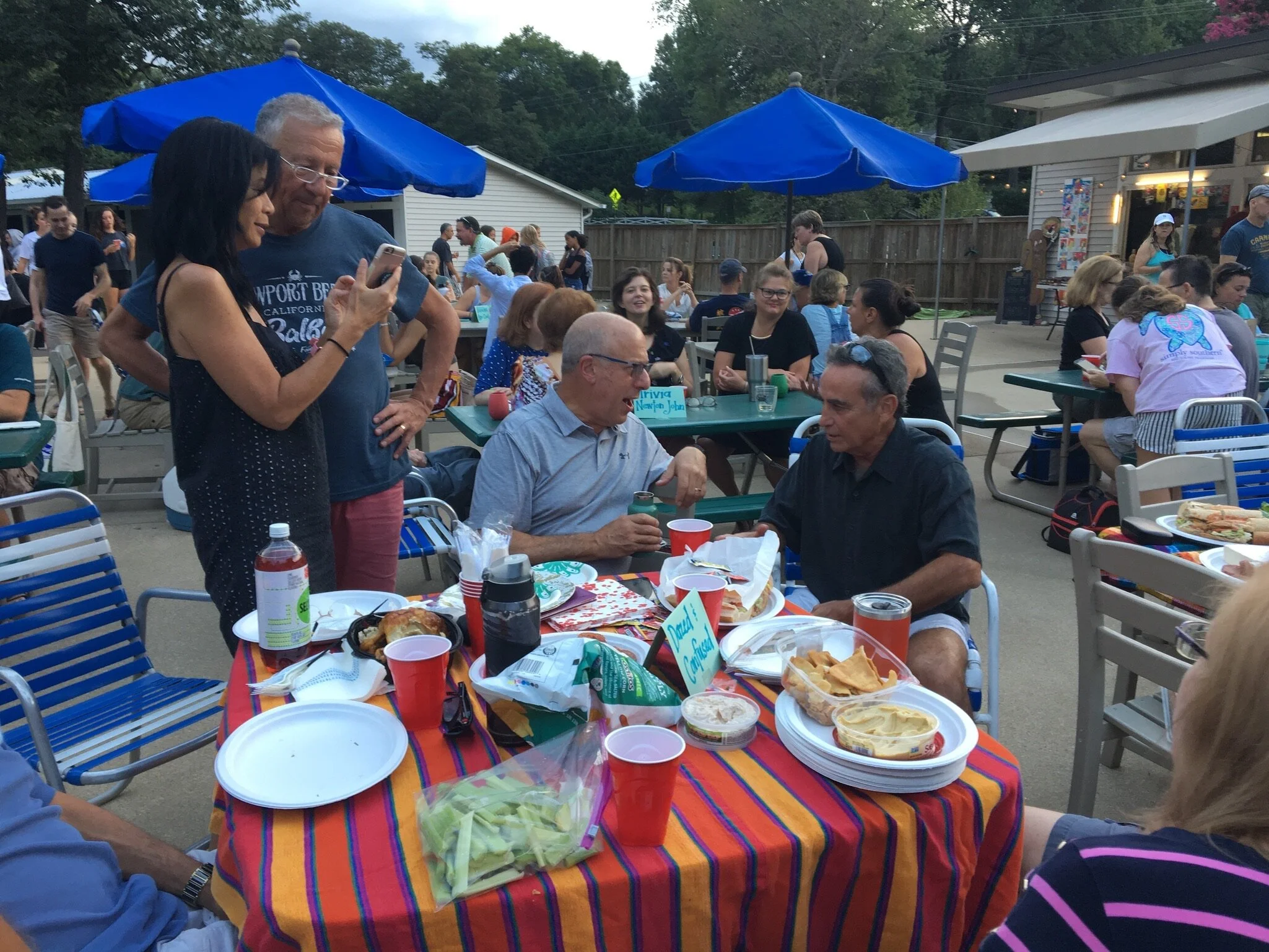 People gathered at an outdoor barbecue, with some talking, eating, and using their phones, under blue umbrellas, in a backyard setting.