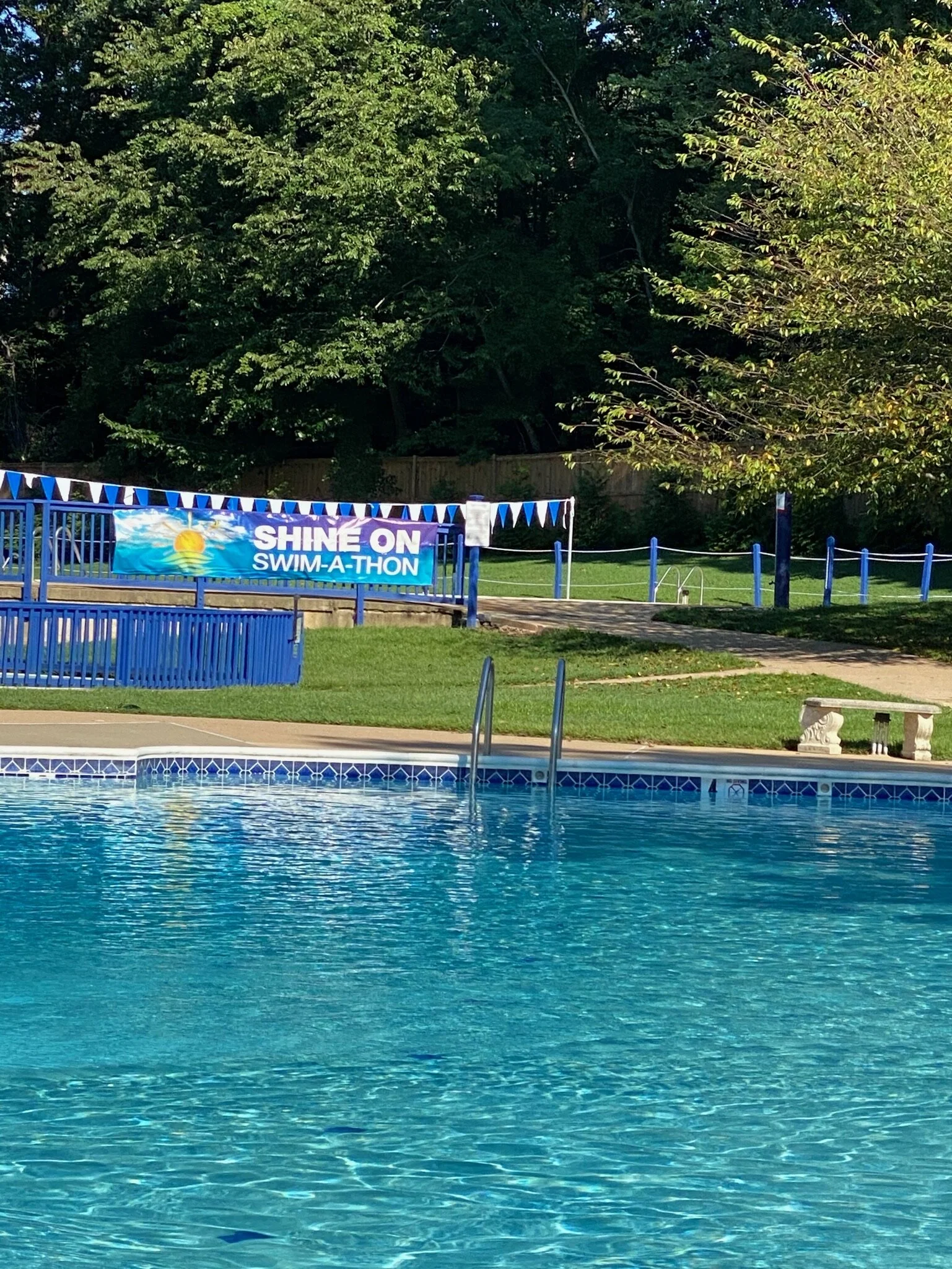 Empty outdoor swimming pool with a blue fence and a 'Shine On Swim-A-Thon' banner, surrounded by green grass, trees, and a pathway.