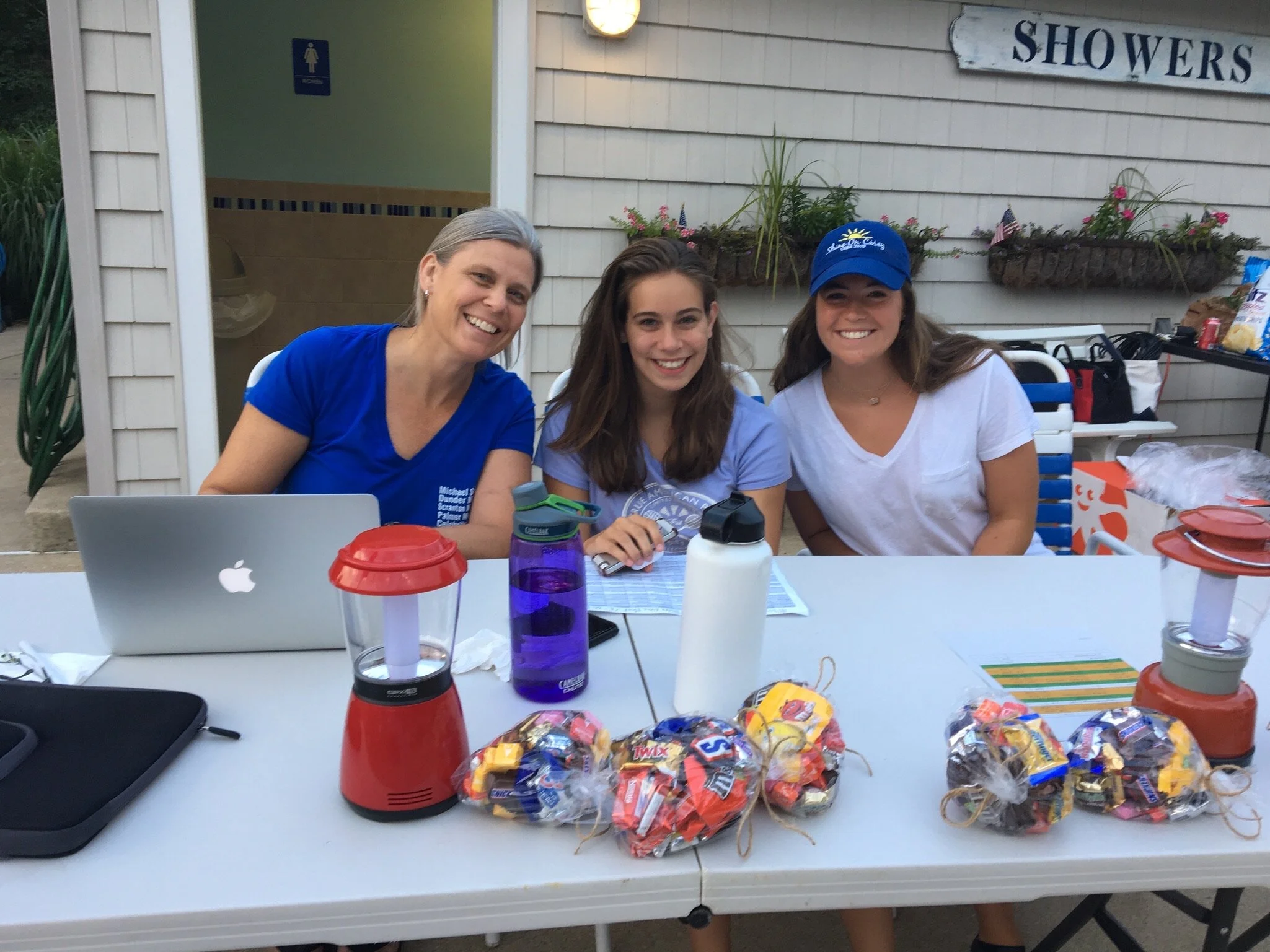 Three women sitting at a table outdoors, smiling for the camera. The table has small lanterns, water bottles, and wrapped snack bags. Behind them is a building with a sign that says 