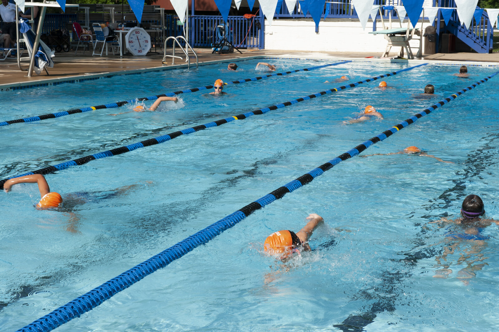 Swimmers in an outdoor swimming pool practicing lap swimming, some wearing orange swim caps.