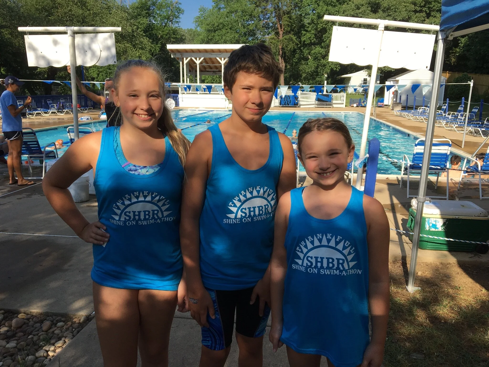 Three children standing by a swimming pool, wearing blue swim team shirts with the initials SHBR, smiling for the camera.