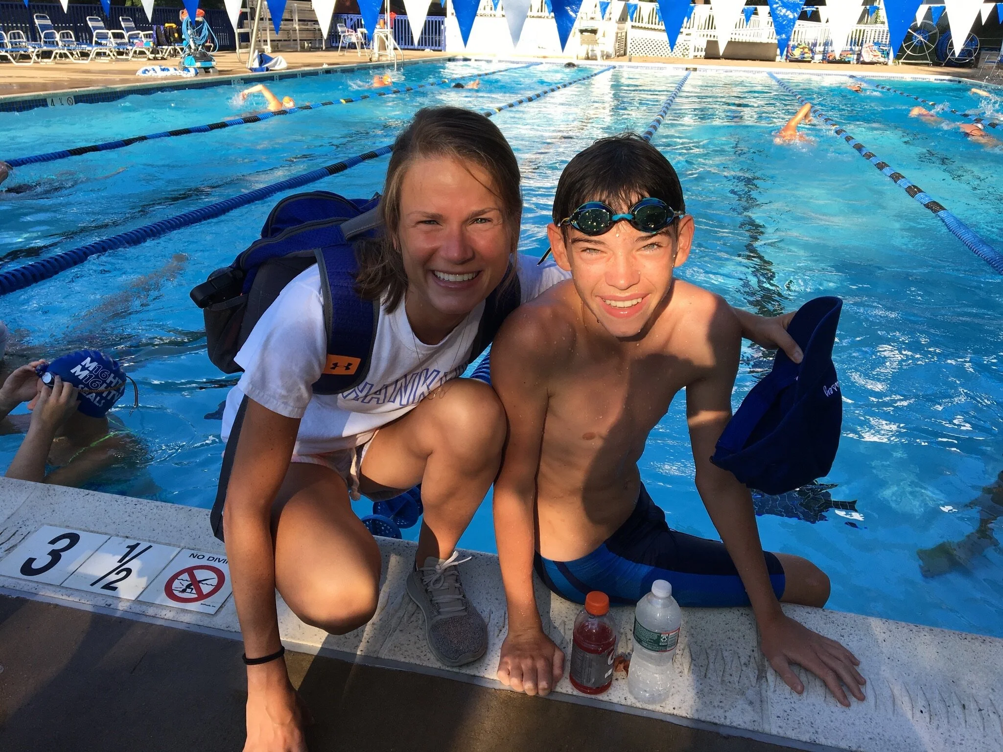 A woman and a young boy are crouching at the edge of an outdoor swimming pool, smiling at the camera. The boy is wearing goggles and swim trunks, while the woman is wearing a white T-shirt, shorts, and a backpack. There are other swimmers in the back