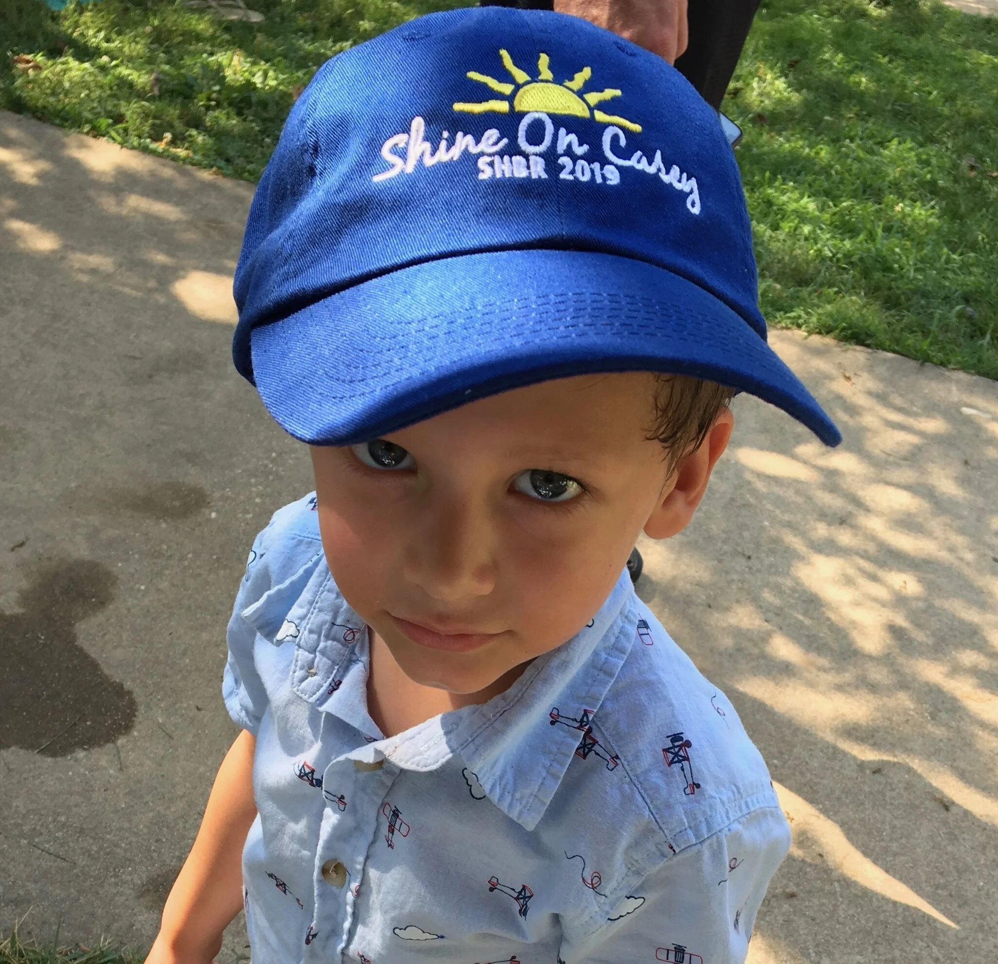 Close-up of a young boy wearing a blue cap with embroidered text and a drawing of a sun, and a light button-up shirt with small airplane and hot air balloon designs, standing outdoors on a paved path with grass and shadows in the background.