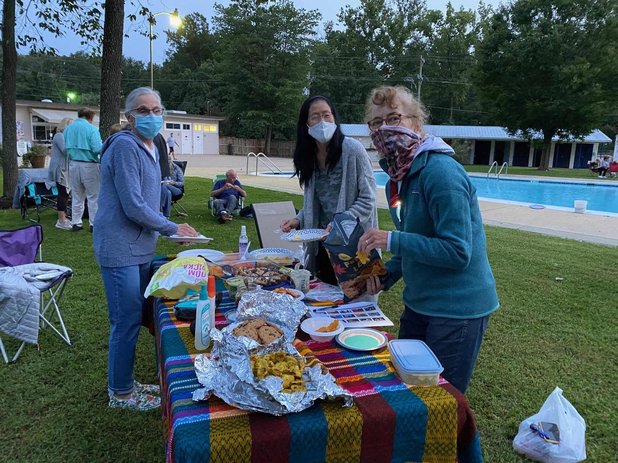 People wearing face masks gathering around a table with food outdoors near a swimming pool in the evening.