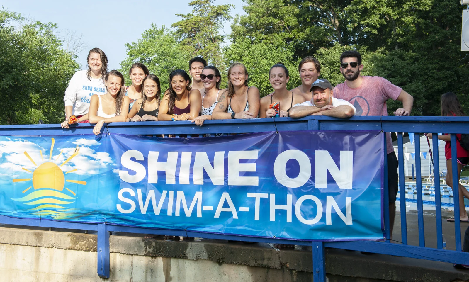 Group of smiling people behind a blue railing at a swim-a-thon event, with a banner that reads "Shine on Swim-a-thon" and a graphic of a shining sun over water, outdoors on a sunny day.