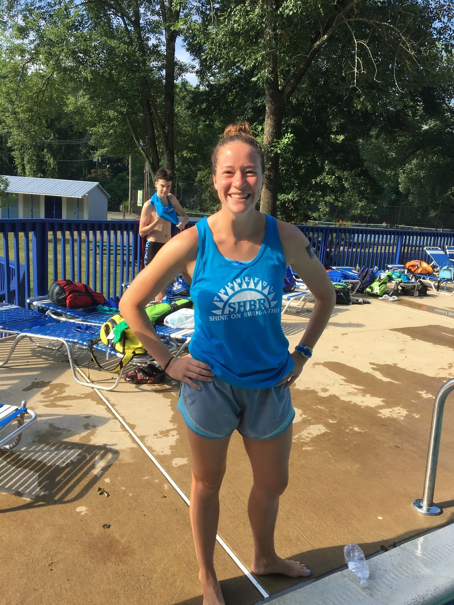 A young woman in a blue swim team shirt and gray shorts standing on a pool deck, smiling with her hands on her hips, with a young boy in swimming gear in the background, and lounge chairs, bags, and trees around.