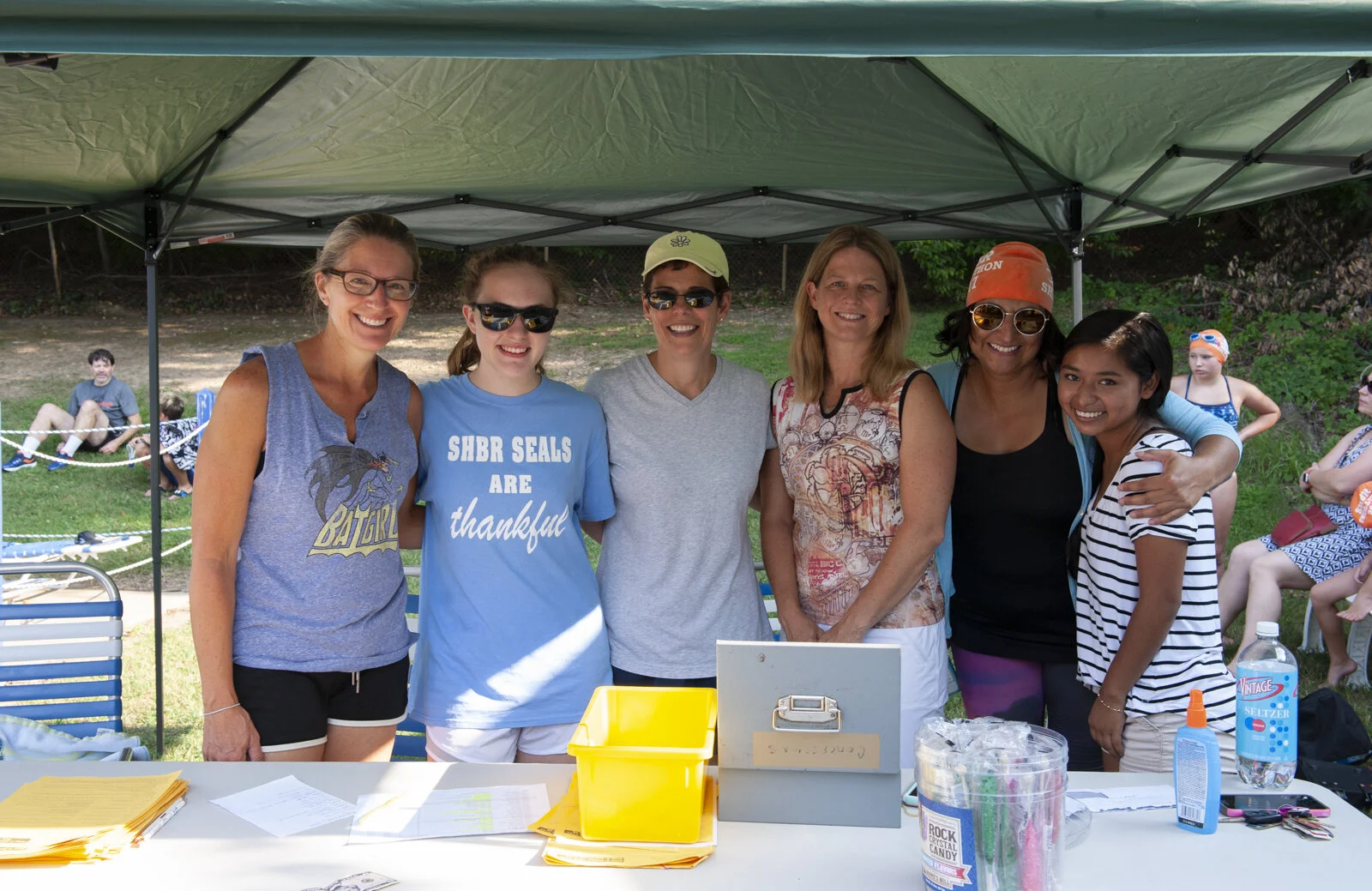 Six women are standing under a green canopy, smiling at the camera, with a table in front of them and children in swimming gear in the background at an outdoor event.
