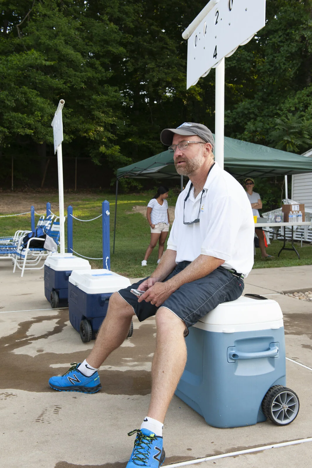 A man with glasses and a beard sitting on a large cooler near a swimming pool. He is wearing a white polo shirt, black shorts, bright blue sneakers, and a dark cap. Behind him, there are two other women near a tent and a table with supplies, and ther