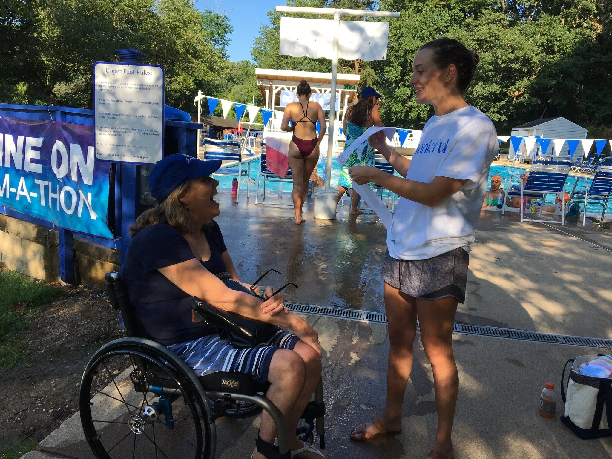 Two women having a conversation near a swimming pool, one sitting in a wheelchair and the other standing. The pool area has chairs, people swimming, and a sign with pool rules in the background.
