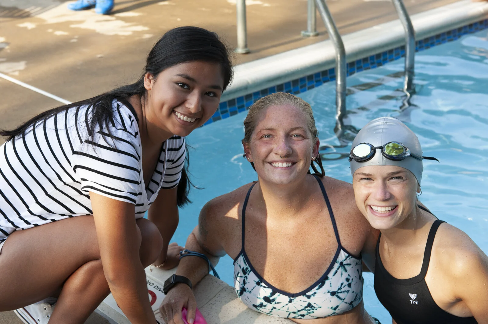 Three women at a swimming pool, smiling and posing for a photo. One woman is sitting on the edge of the pool wearing a black swimsuit, the other two are standing in the pool, one wearing a striped swimsuit and the other wearing a black swimsuit with 