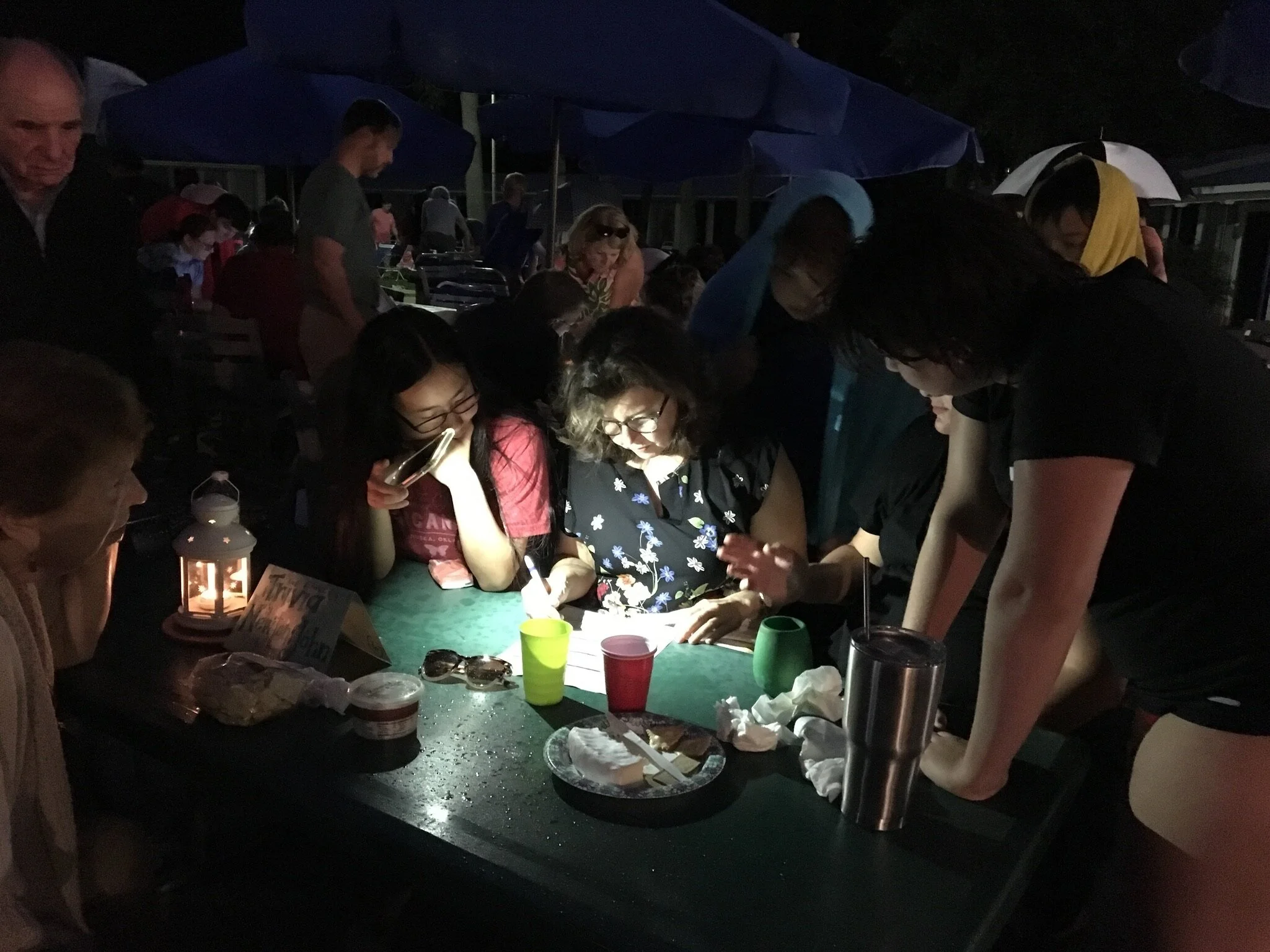 A dark outdoor gathering with people around a table, illuminated by a small lantern, some looking at a book or photo, with umbrellas and other groups visible in the background.