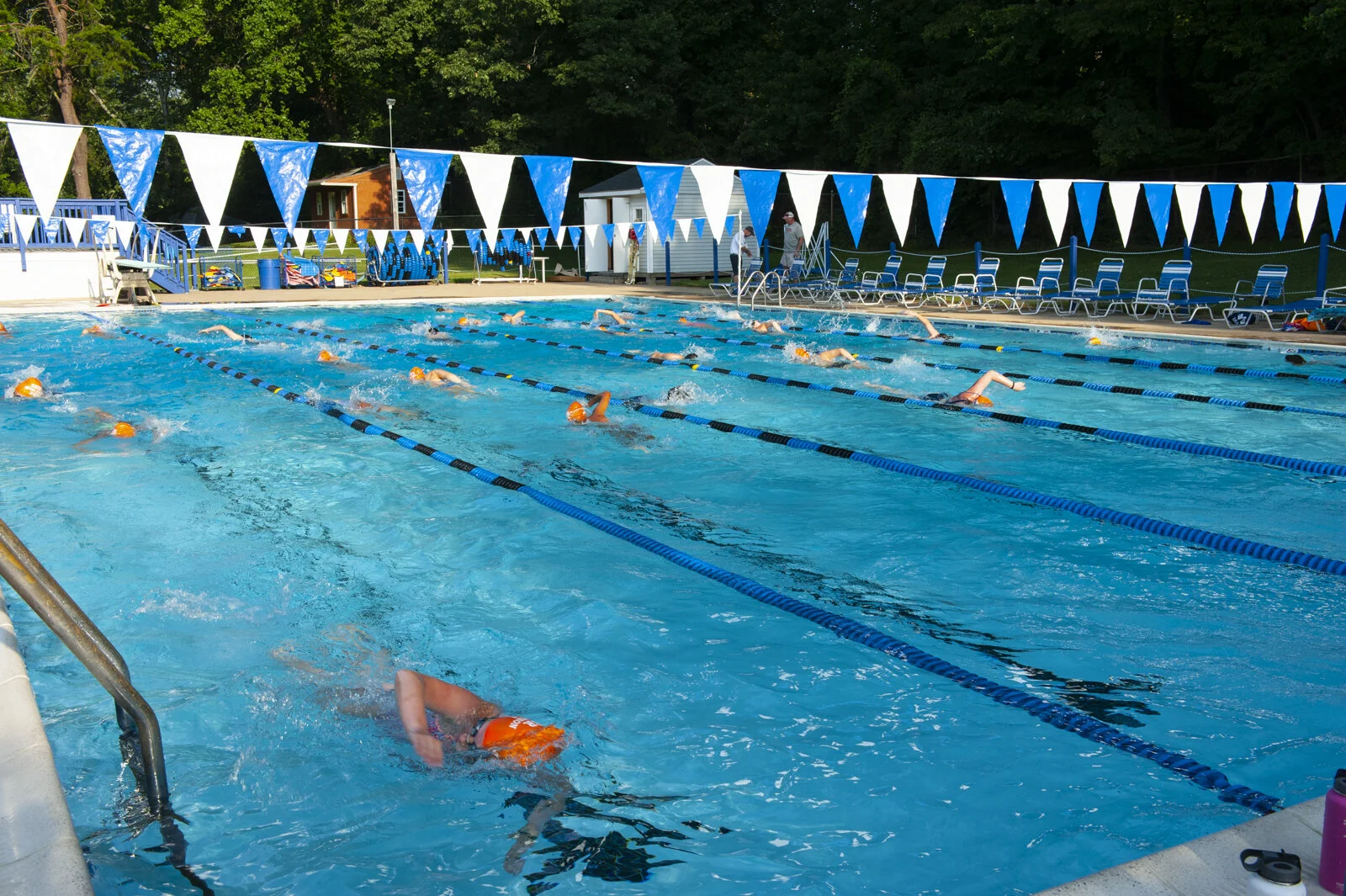Swimmers swimming laps in an outdoor swimming pool, with pool lanes marked by blue and black lane dividers, and flags hanging above the pool, surrounded by trees and lounge chairs.