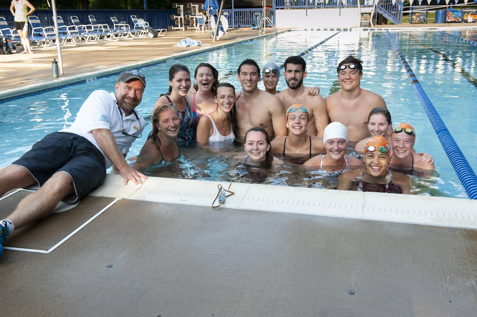A group of people, including swim instructors and swimmers, posing together in a swimming pool, some wearing swim caps and goggles, smiling for a group photo.