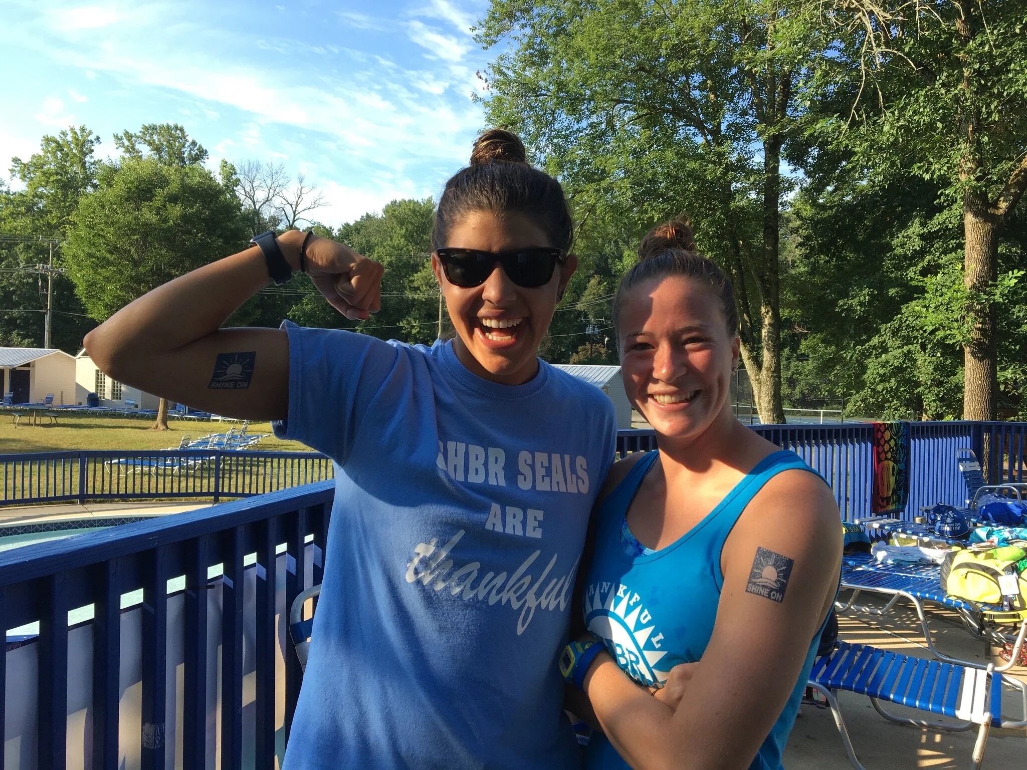 Two women wearing blue tank tops with event logos, smiling outdoors near a swimming pool, with trees and sunlit sky in the background.