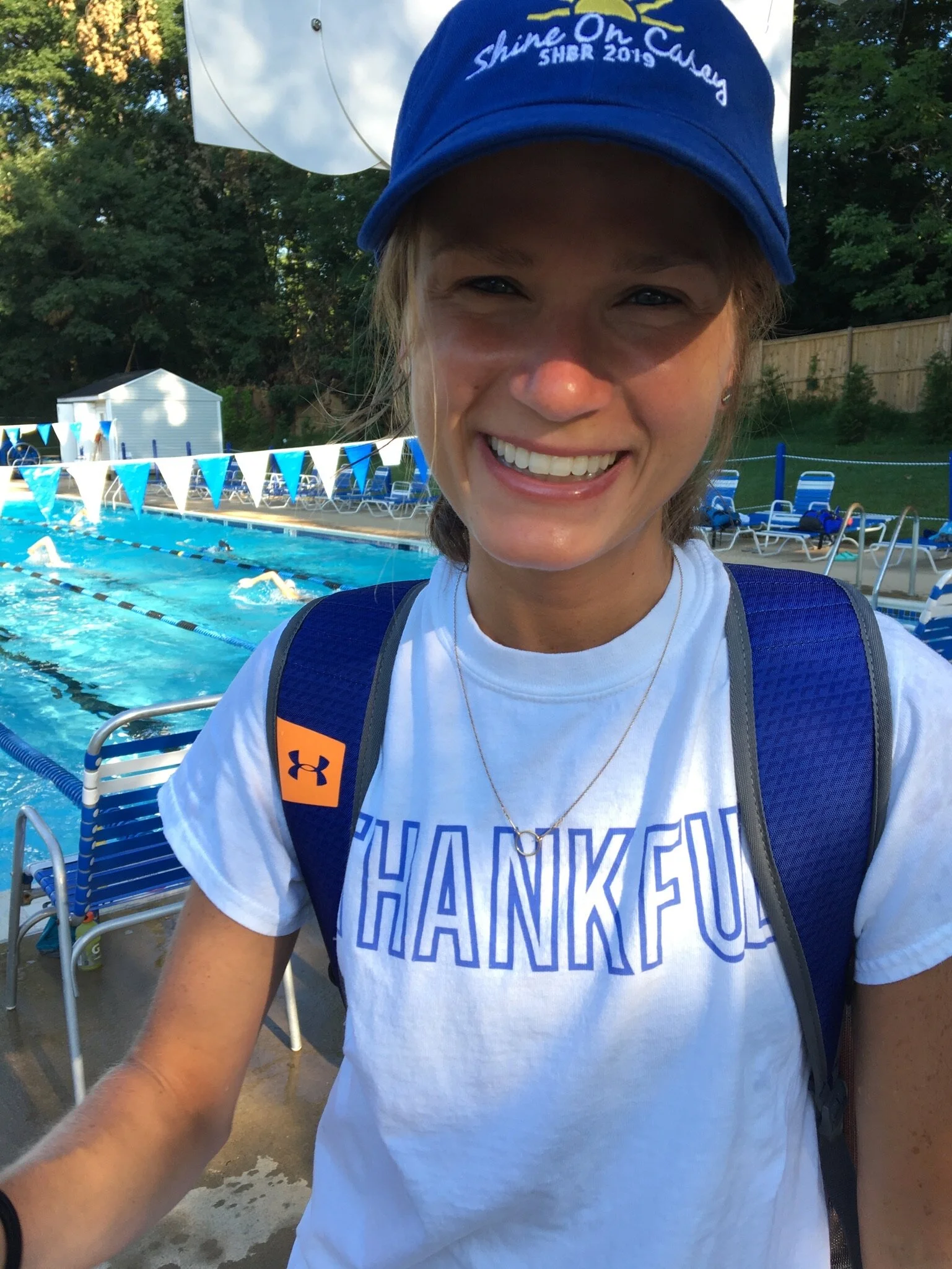Smiling young woman at a swimming pool, wearing a blue cap with text, white t-shirt with word 'thankful', and a blue backpack.