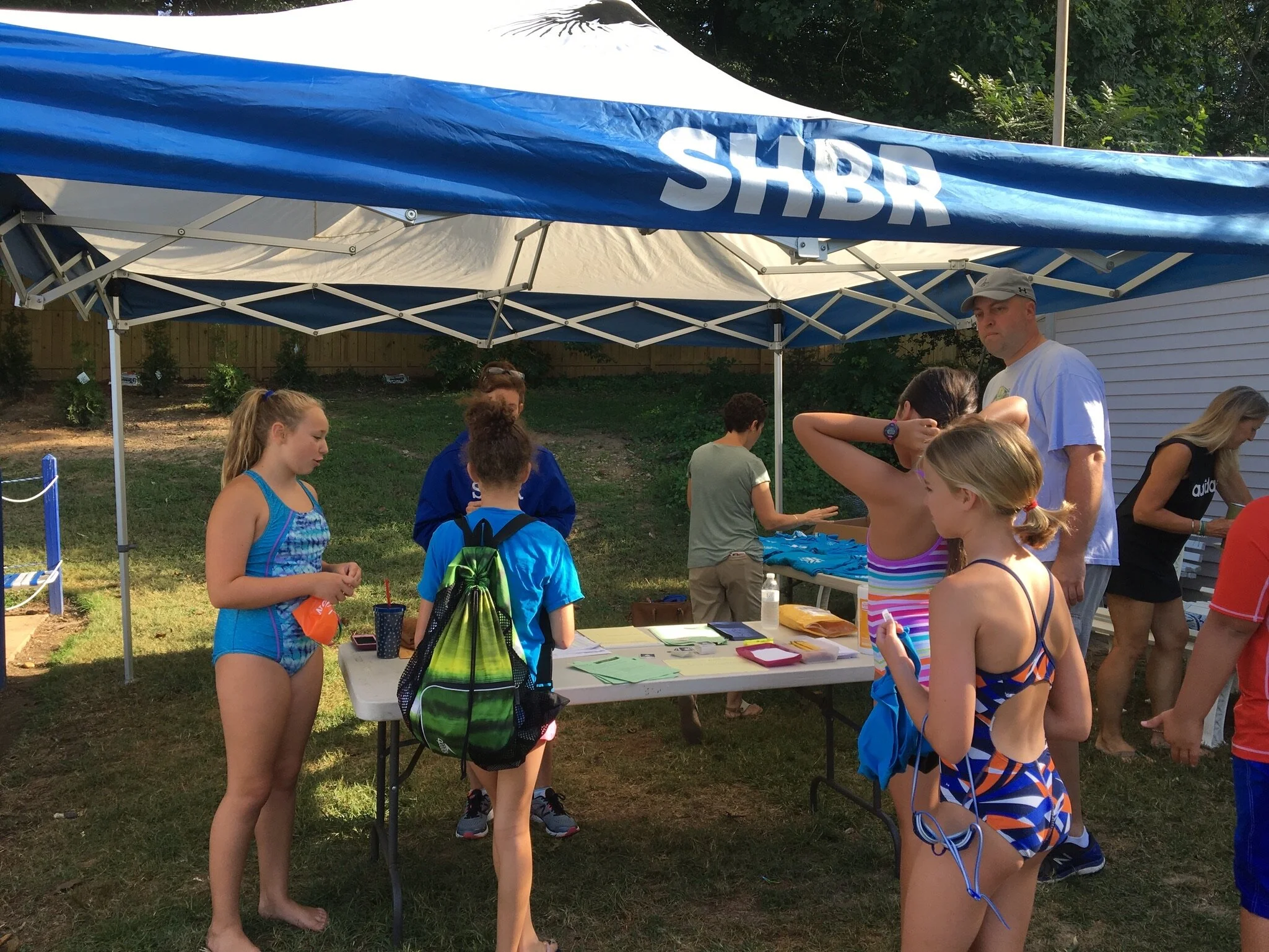 Children in swimsuits standing and waiting in line near registration table under a blue and white canopy tent with 'SHBR' printed on it, outdoors on grass and dirt, with some trees and a white building in the background.