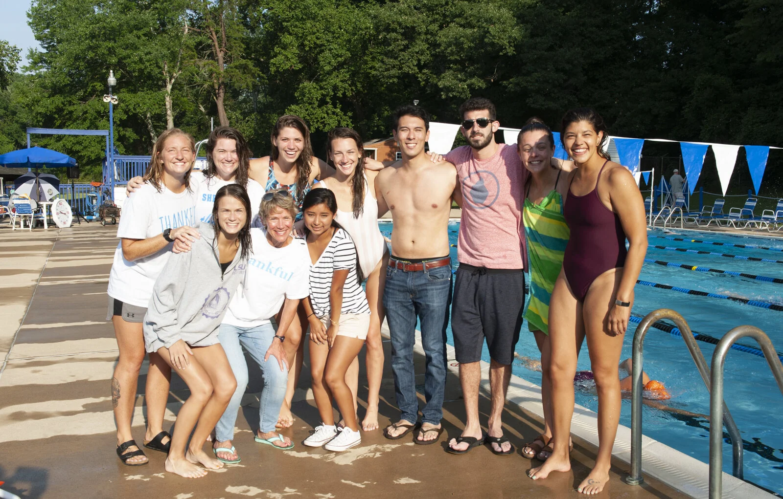 A group of thirteen people, including men and women of various ages, posing together by an outdoor swimming pool with trees in the background. Some are in swimsuits, others in casual clothes, smiling and standing close to each other.