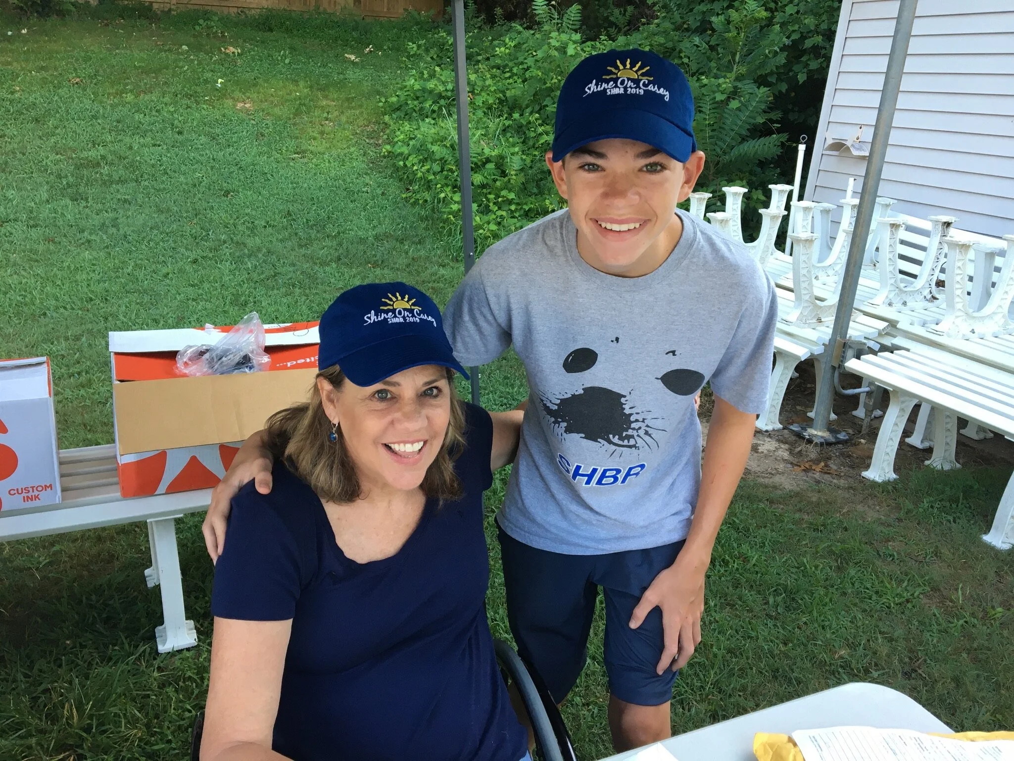 A woman and a teenage boy smiling outdoors. Both are wearing navy blue caps with gold crown logos and the text ‘Shine On Casey 2019’. The woman is seated, wearing a navy blue shirt, and the boy is standing beside her, wearing a gray T-shirt with a wo