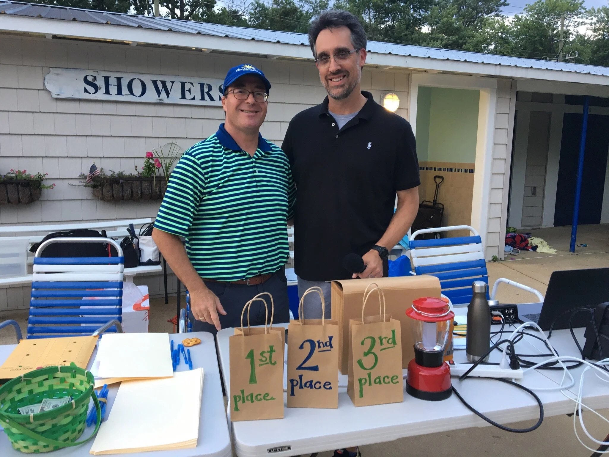 Two men standing behind a table with first, second, and third place awards at an outdoor event near a shower house.