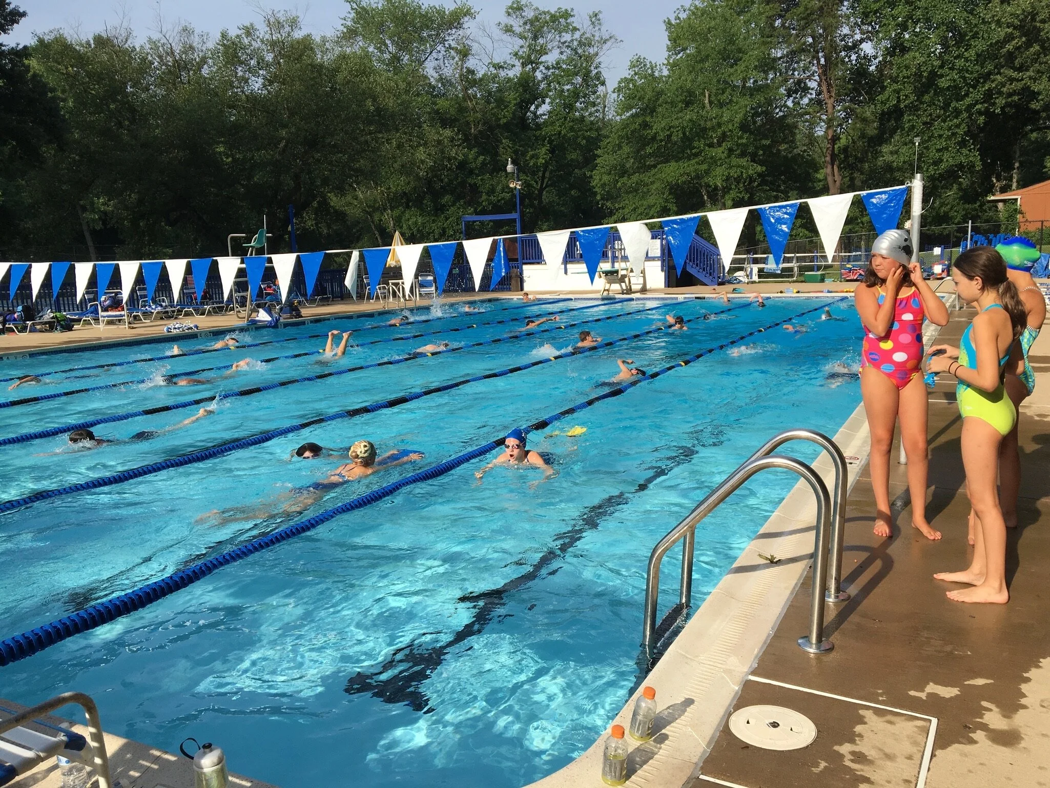 Children swimming and practicing laps in an outdoor swimming pool with blue lane dividers, while two girls in colorful swimsuits and swim caps stand at the poolside.