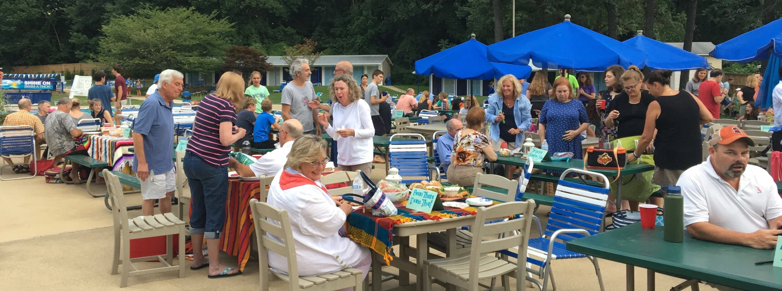 People gathered at an outdoor event with decorated tables, blue umbrellas, and trees in the background, enjoying food and socializing.