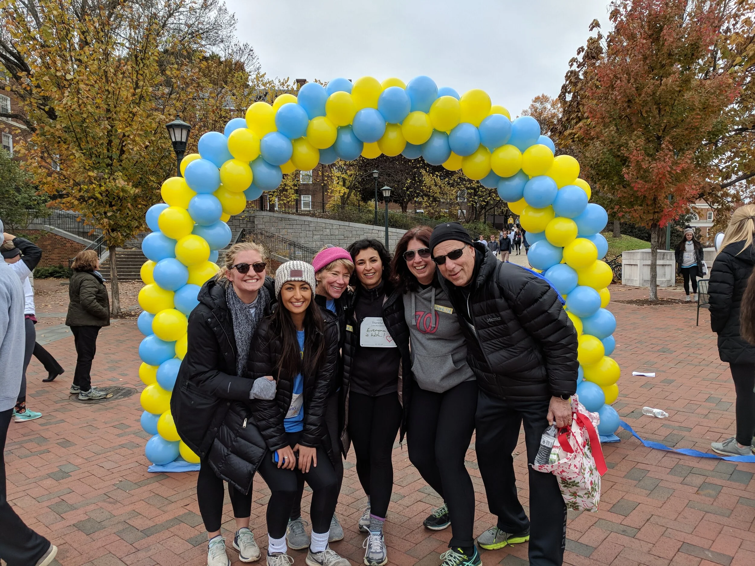 Group of six people standing under a yellow and blue balloon arch at an outdoor event on a cloudy day, with autumn trees and other participants in the background.