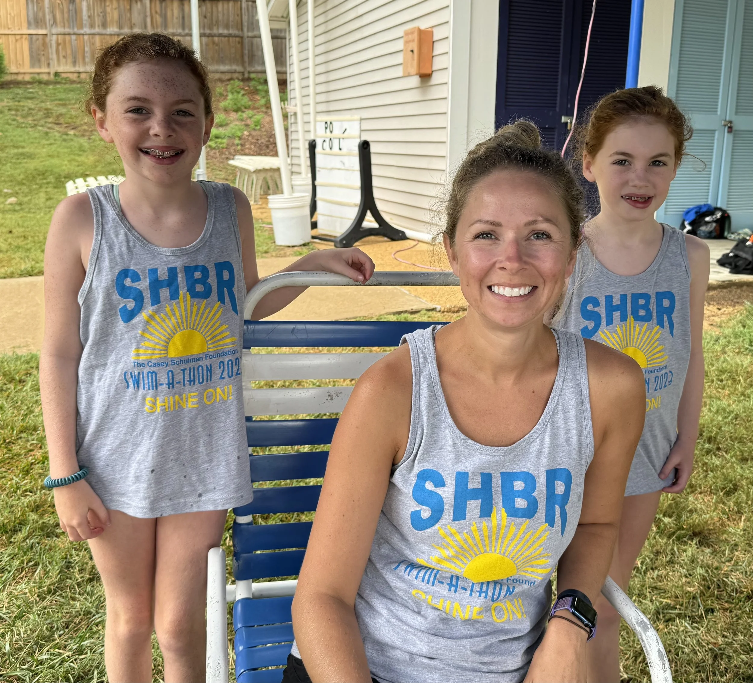 A woman and two girls outdoors, all wearing matching gray tank tops with blue and yellow lettering for a swimathon event, smiling at the camera.