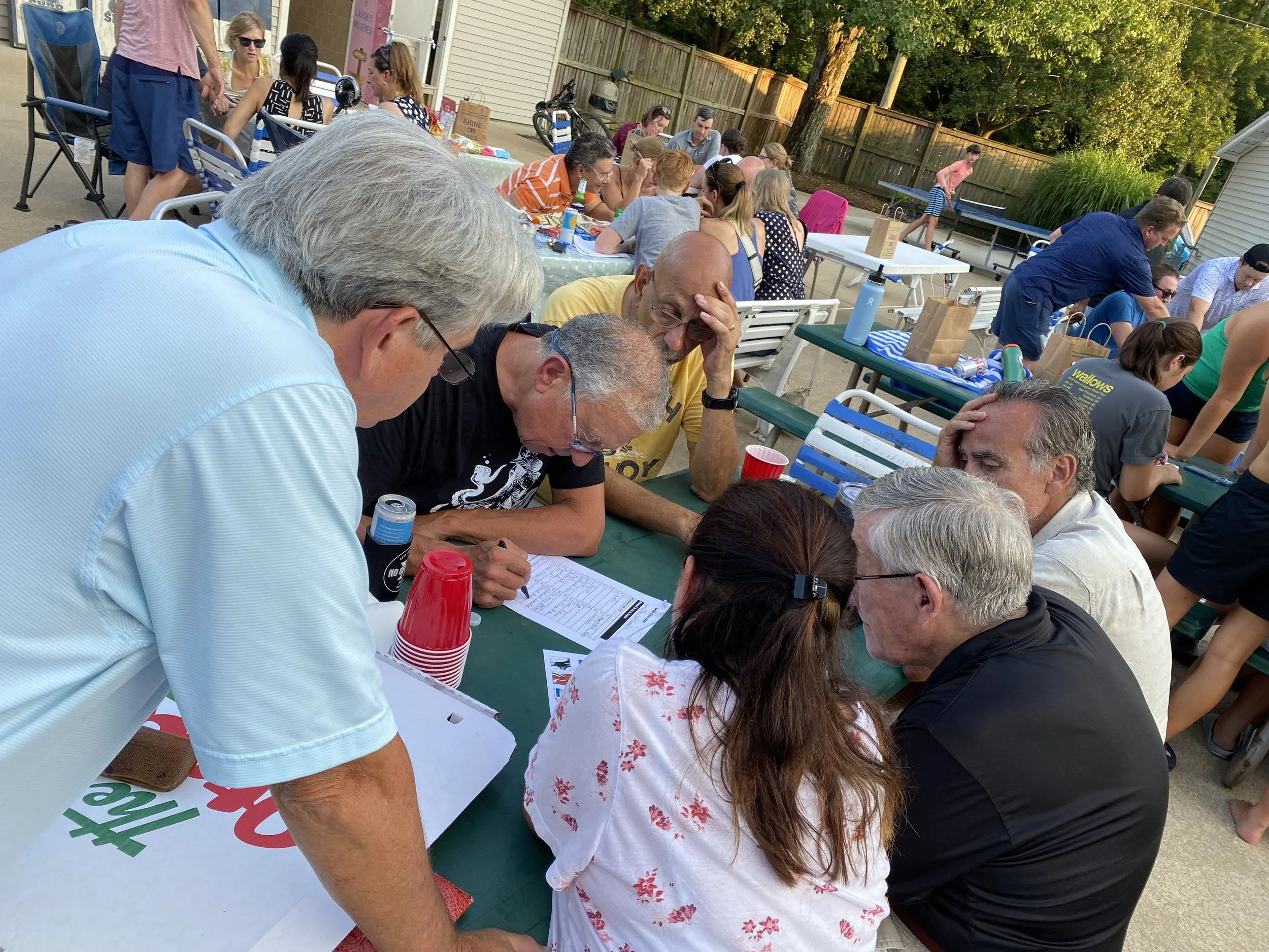 A group of people sitting and standing around a table outdoors, engaged in a discussion. There are many tables and chairs, with more people in the background, and trees surrounding the area.