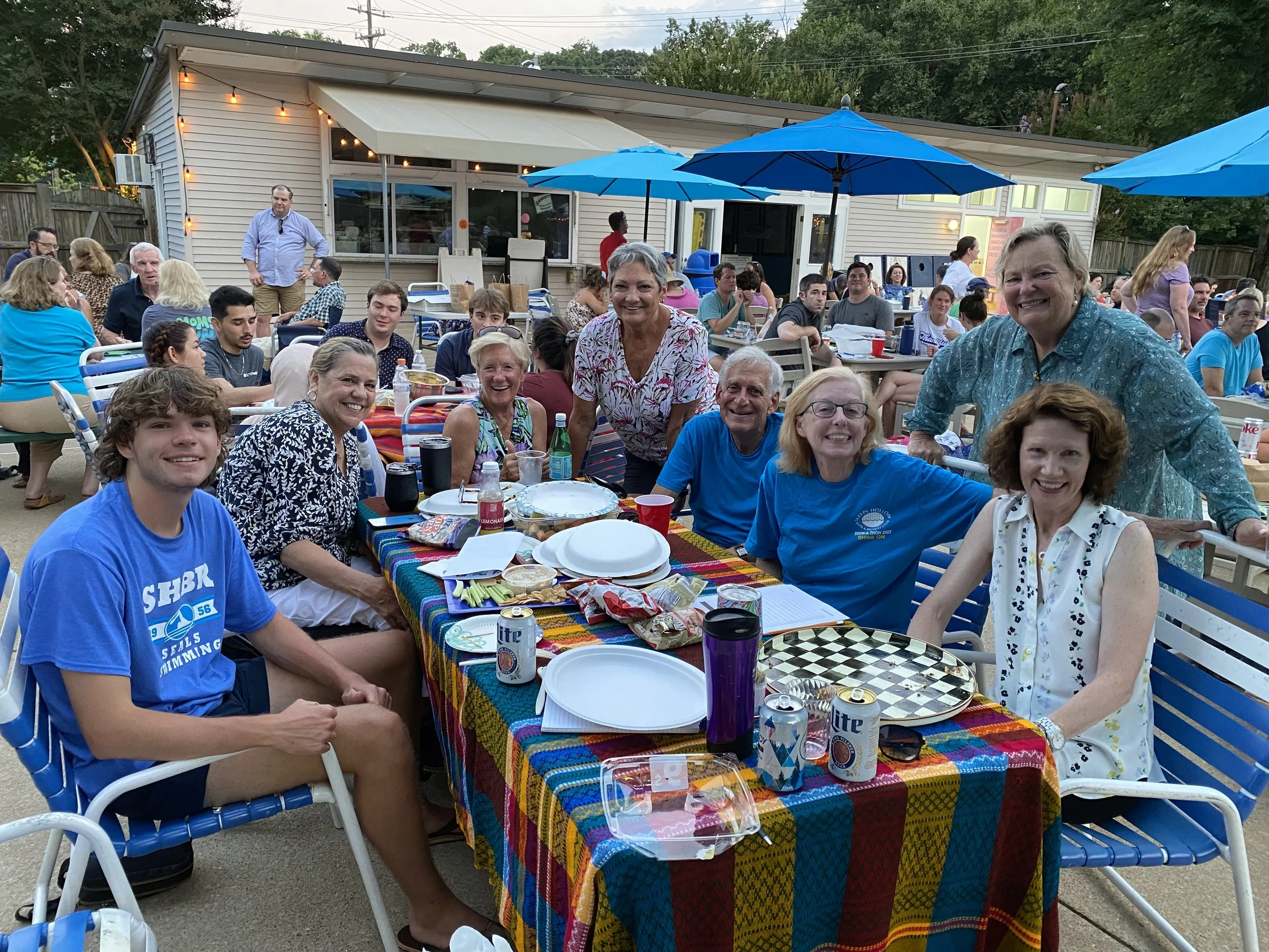 Group of people gathered around a table outdoors at a party or celebration, with many smiling and enjoying food and drinks.