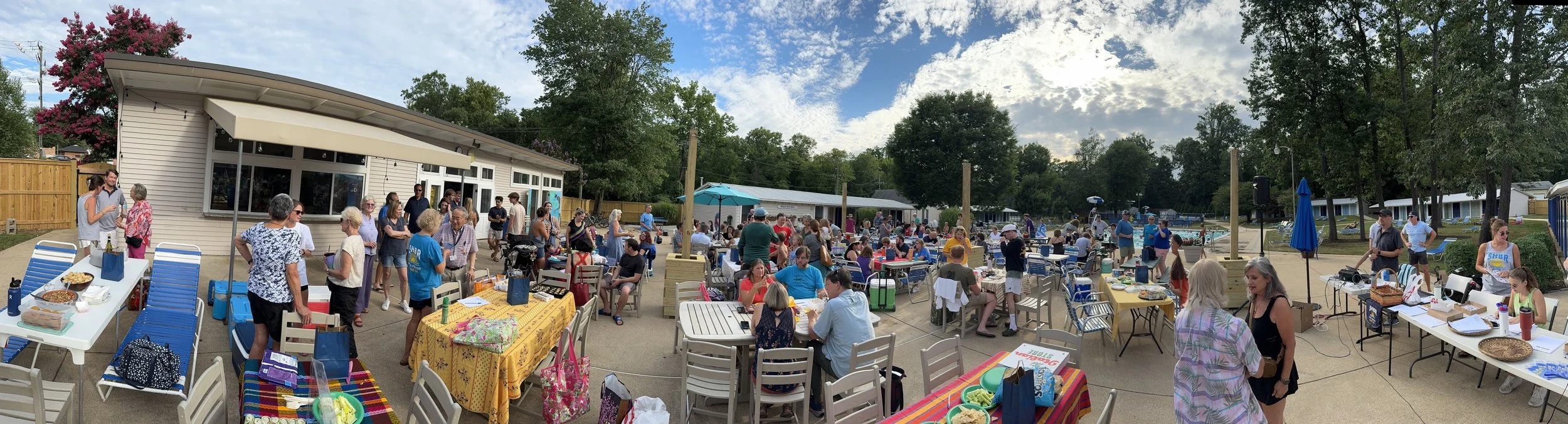 A large outdoor gathering with many people seated at tables and socializing, event setup near a pool with trees and buildings in the background, and food on tables.