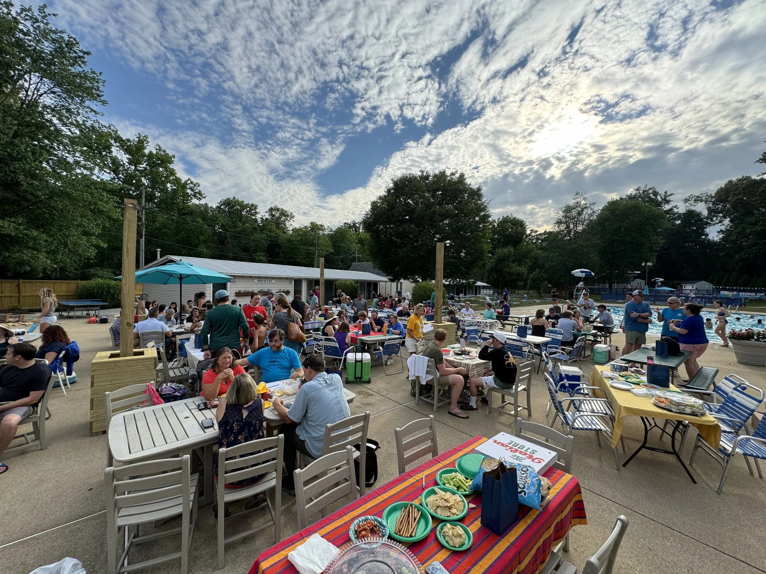 People gathered at an outdoor poolside party with tables, food, and umbrellas on a sunny day with a partly cloudy sky.
