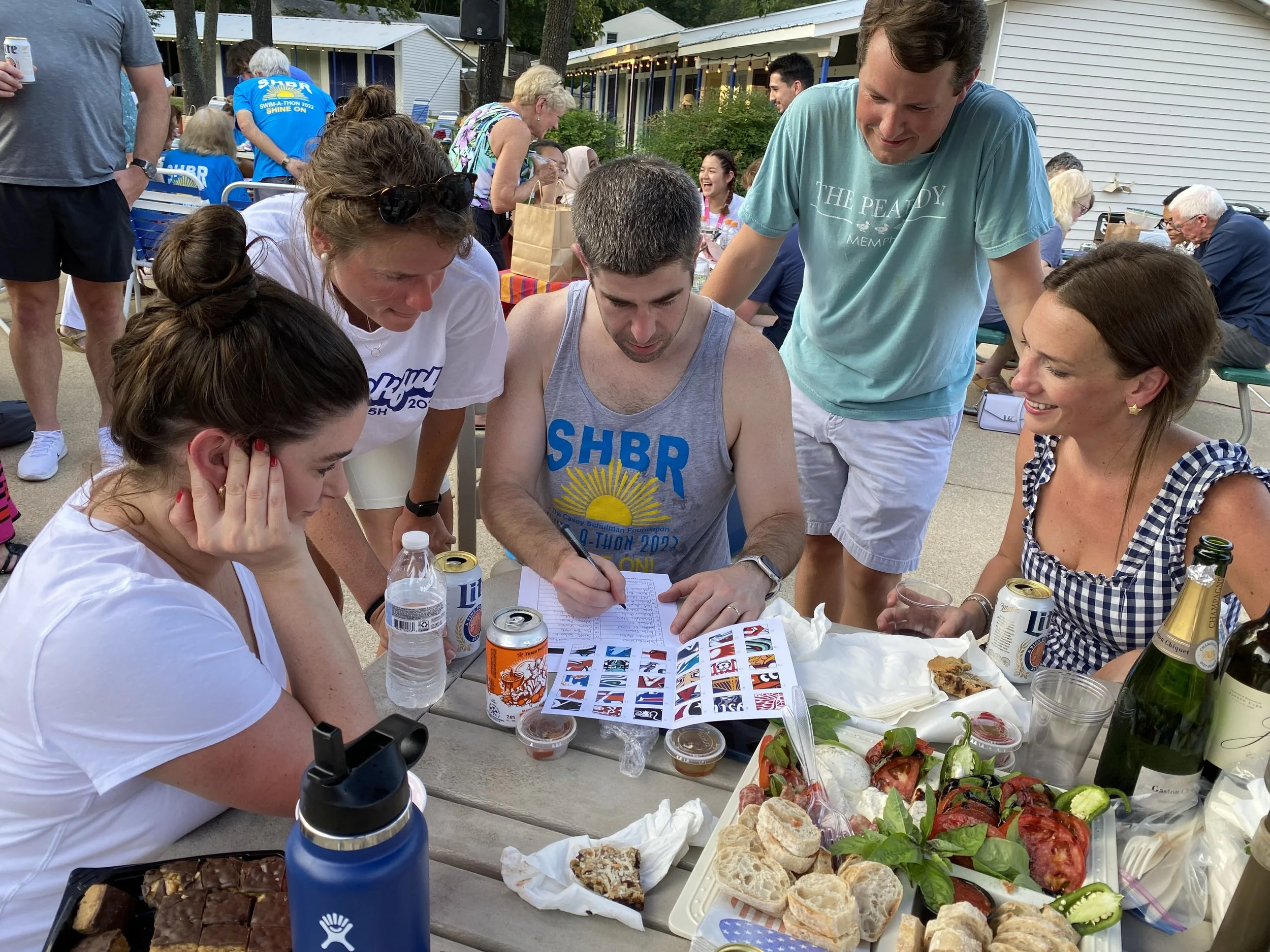 Group of people gathered around a table at an outdoor party, signing a book, with food and drinks on the table, some people in the background, and a house in the distance.