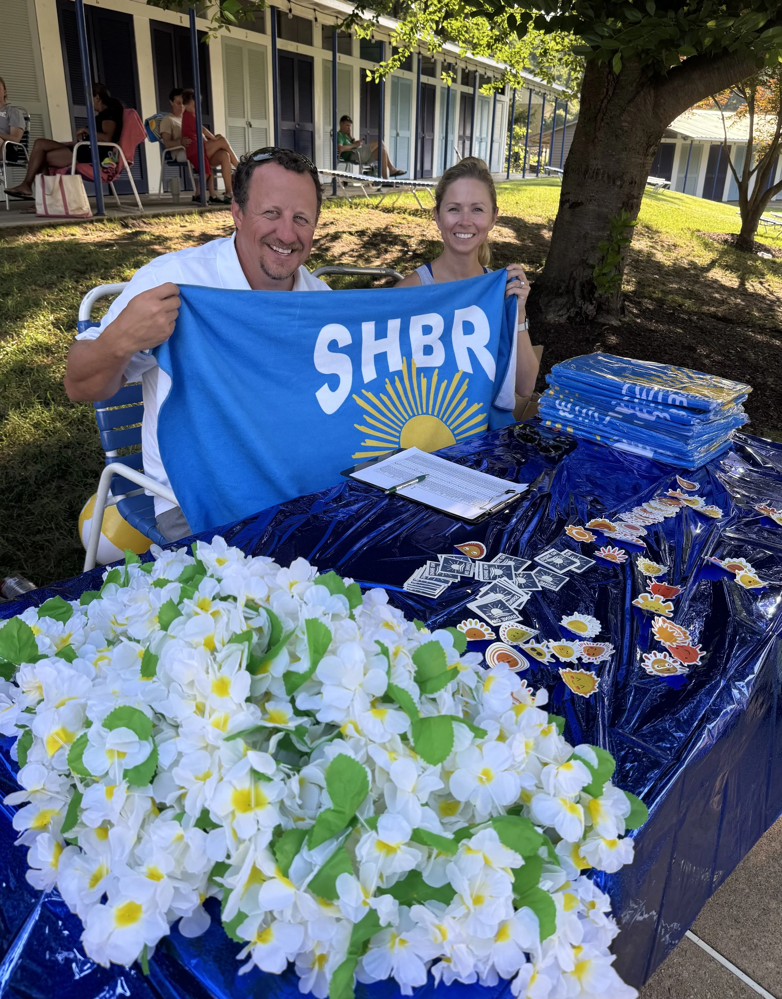 Two smiling people sitting at a table outdoors holding a blue SHBR flag. The table has a dark blue tablecloth, stickers, a clipboard, and folded blue items. There are white and yellow artificial flowers in the foreground and other people sitting on c