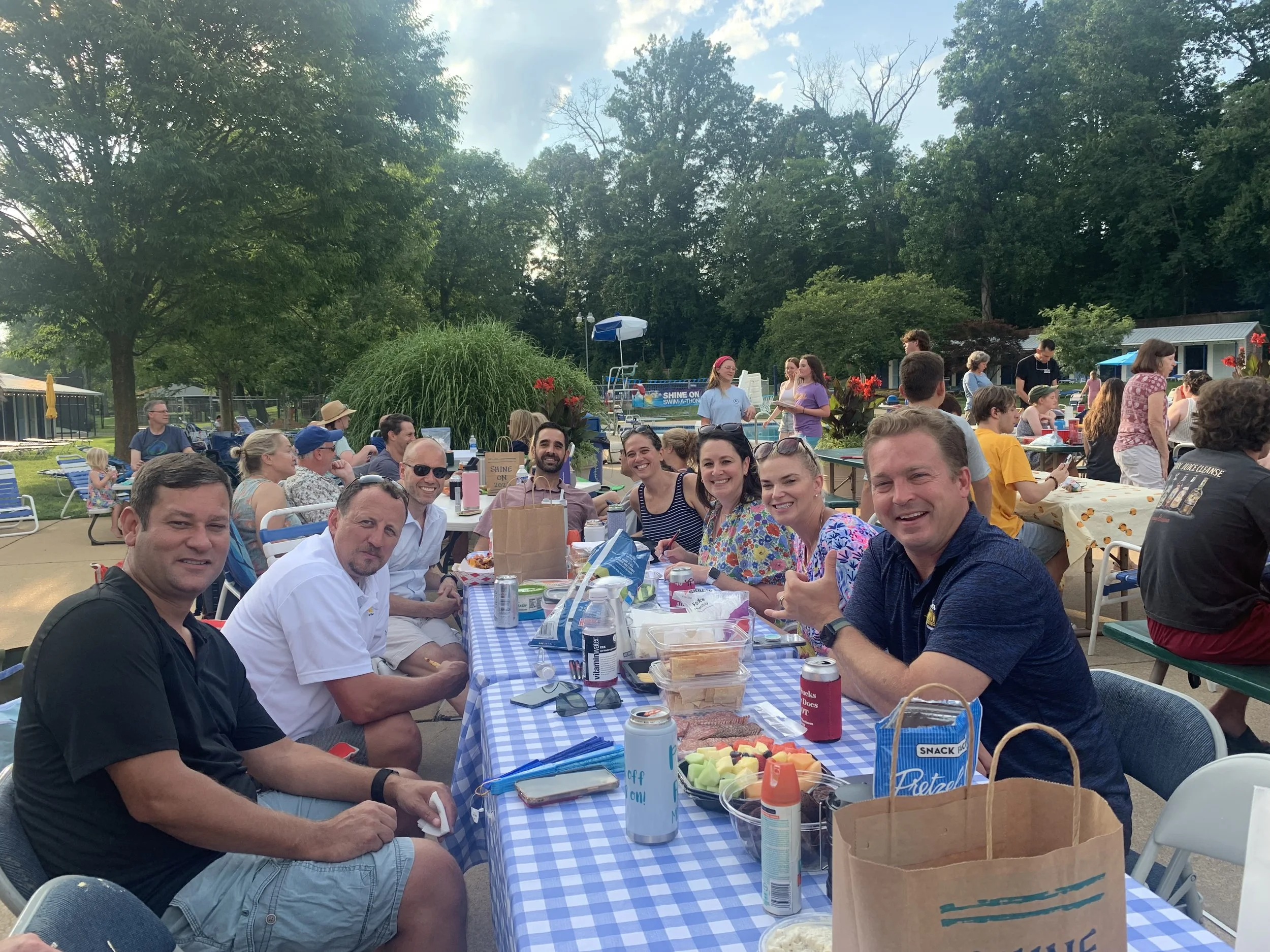 Group of people at an outdoor gathering, sitting at a table with food and drinks, smiling and socializing, with trees and more people in the background.