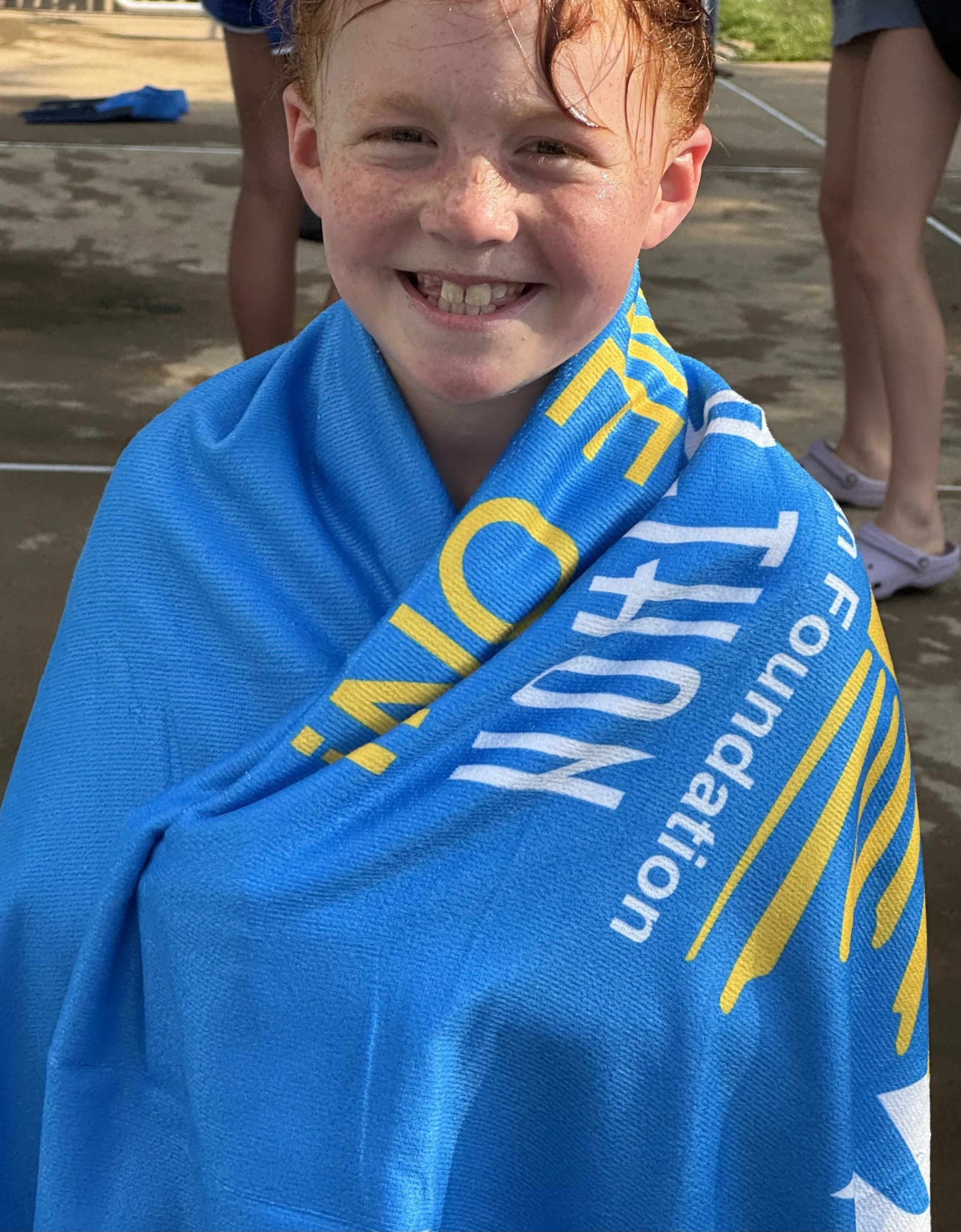 Young boy with red hair and freckles wrapped in a blue towel, smiling at the camera, outdoors on a concrete surface.