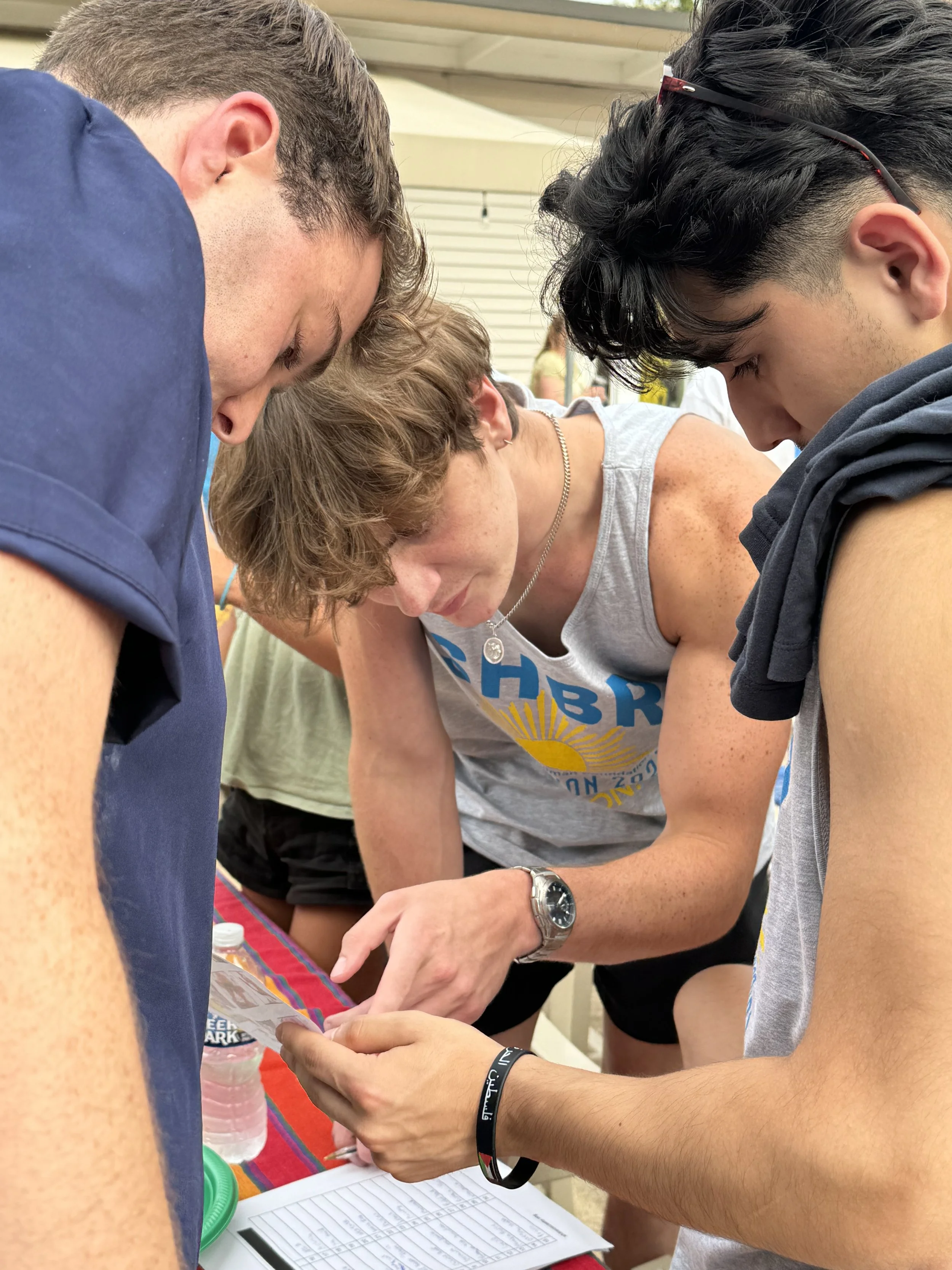 Three young men gathered around a table, looking intently at a phone held by one of them. The scene appears to be at an outdoor event or gathering under a covered area, with a water bottle and a notebook on the table.