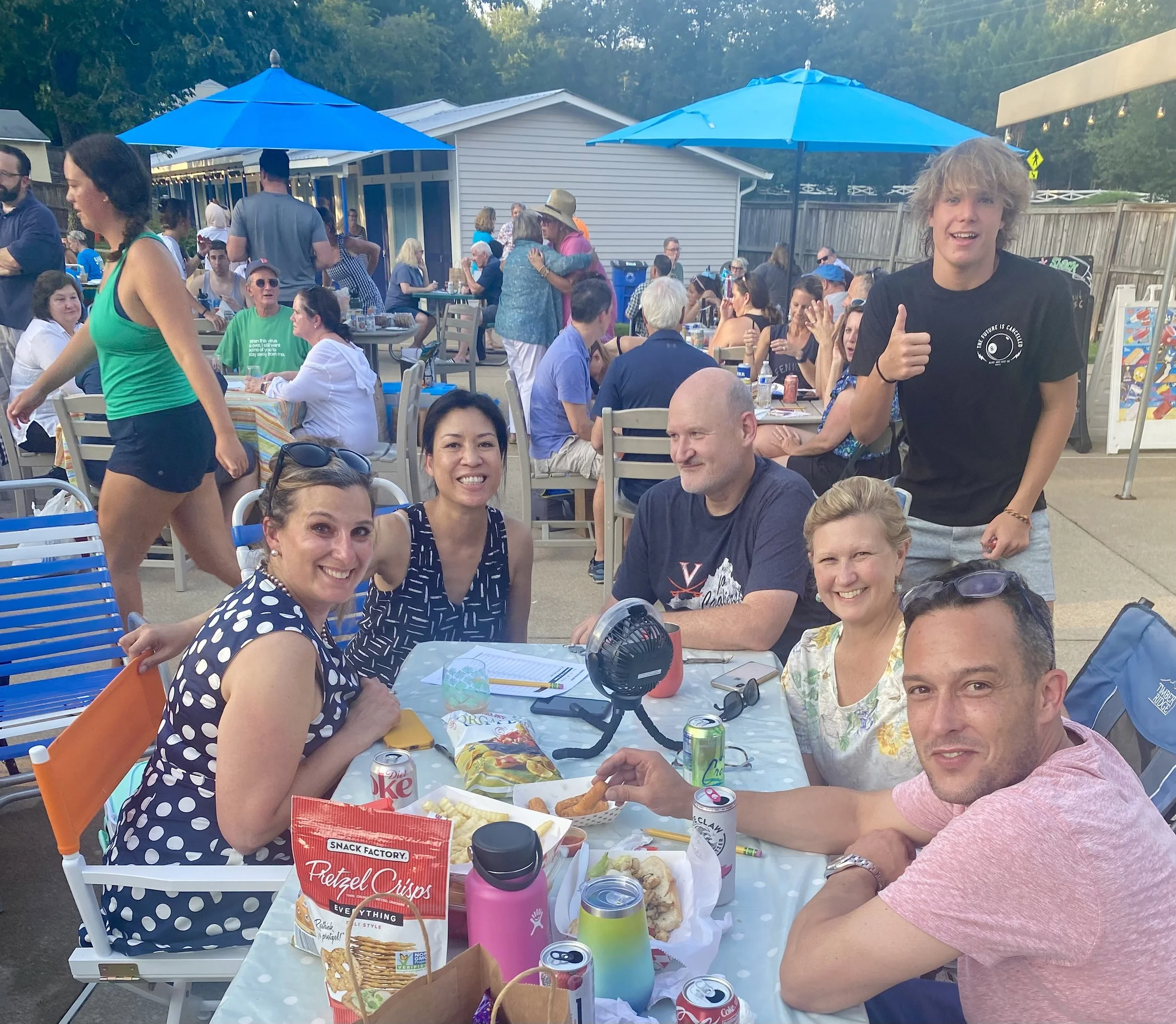 A group of six people sitting around a table with food and drinks at an outdoor gathering, with many other people in the background enjoying the event.
