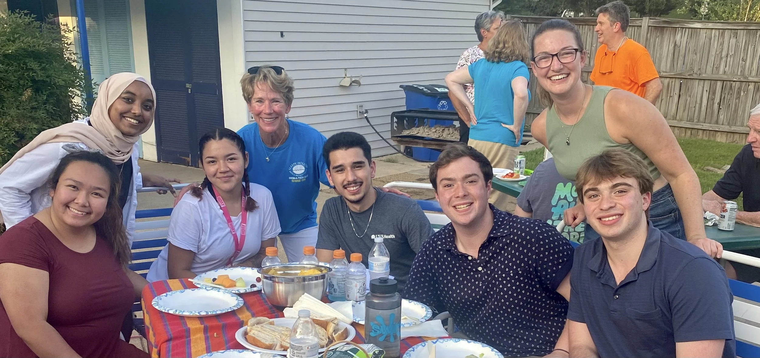 A diverse group of people at a backyard gathering, smiling and enjoying food around a table with plates, drinks, and sandwiches, with some standing and some sitting, in a suburban backyard with a fence and house in the background.