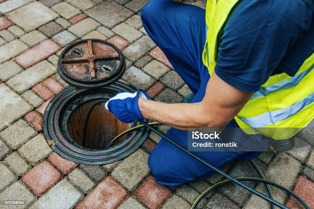 A worker in a yellow safety vest and blue uniform kneeling on a brick sidewalk, using tools to perform maintenance or repair on a sewer or drain cover.