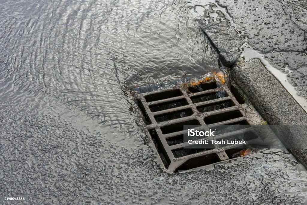 A storm drain on a street surrounded by puddles with water flowing over the surface.