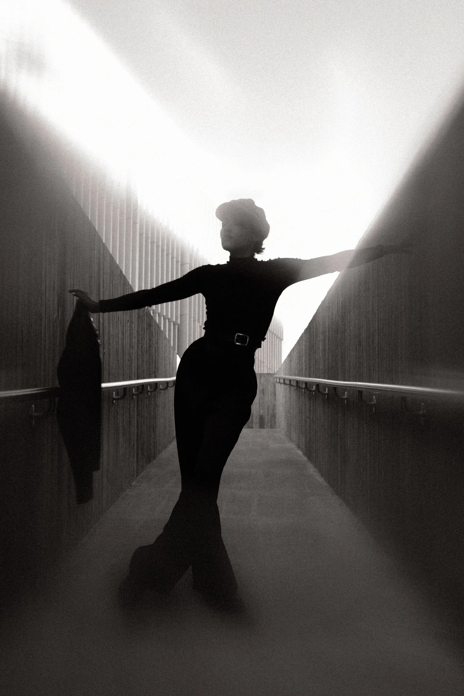 Silhouette of a woman standing in a narrow hallway with her arms extended, wearing a hat and dark clothing, with high contrast lighting creating a dramatic effect.