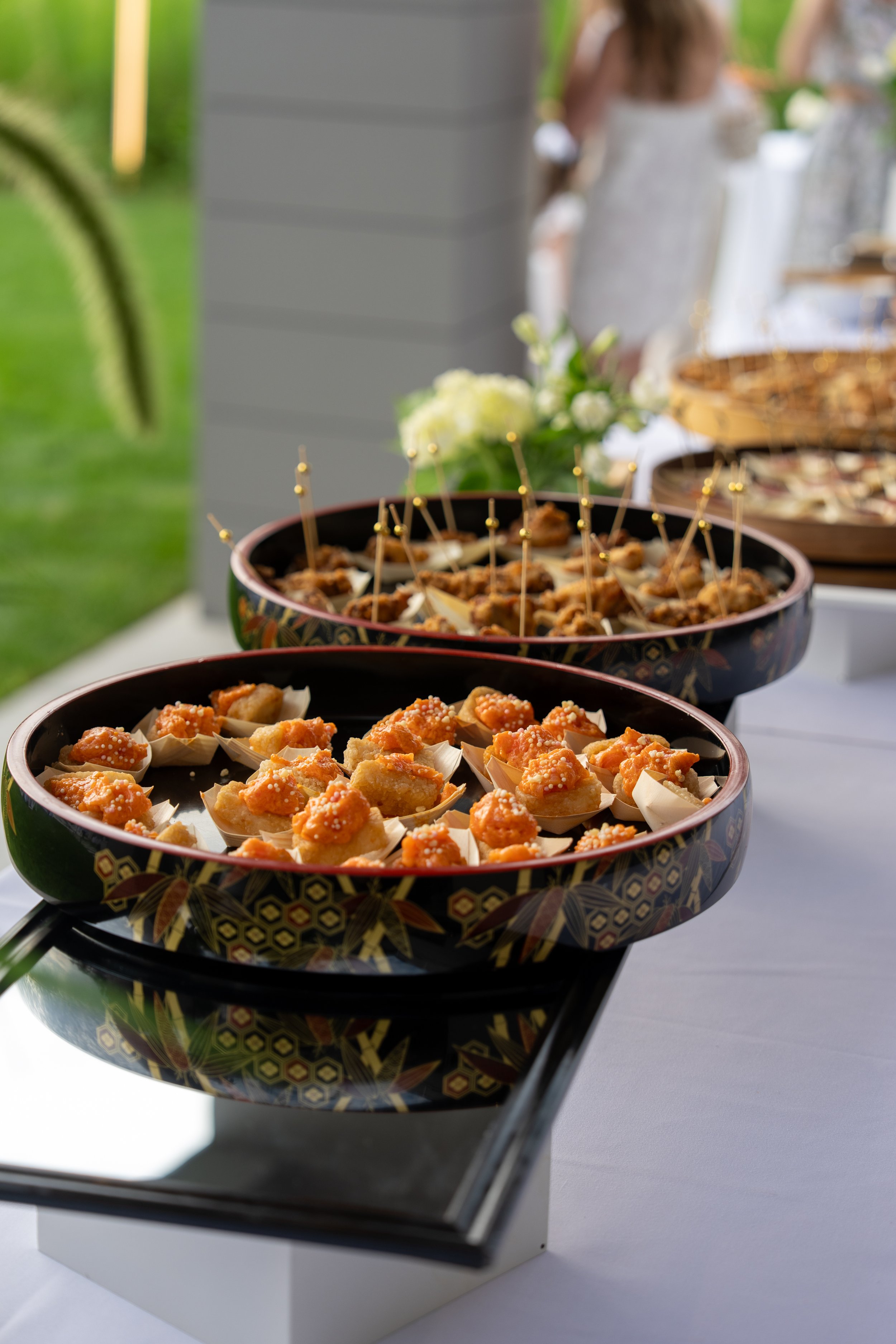 Close-up of black decorative bowls filled with appetizers on a table at a catered event, with a blurred background including a person in a white dress.