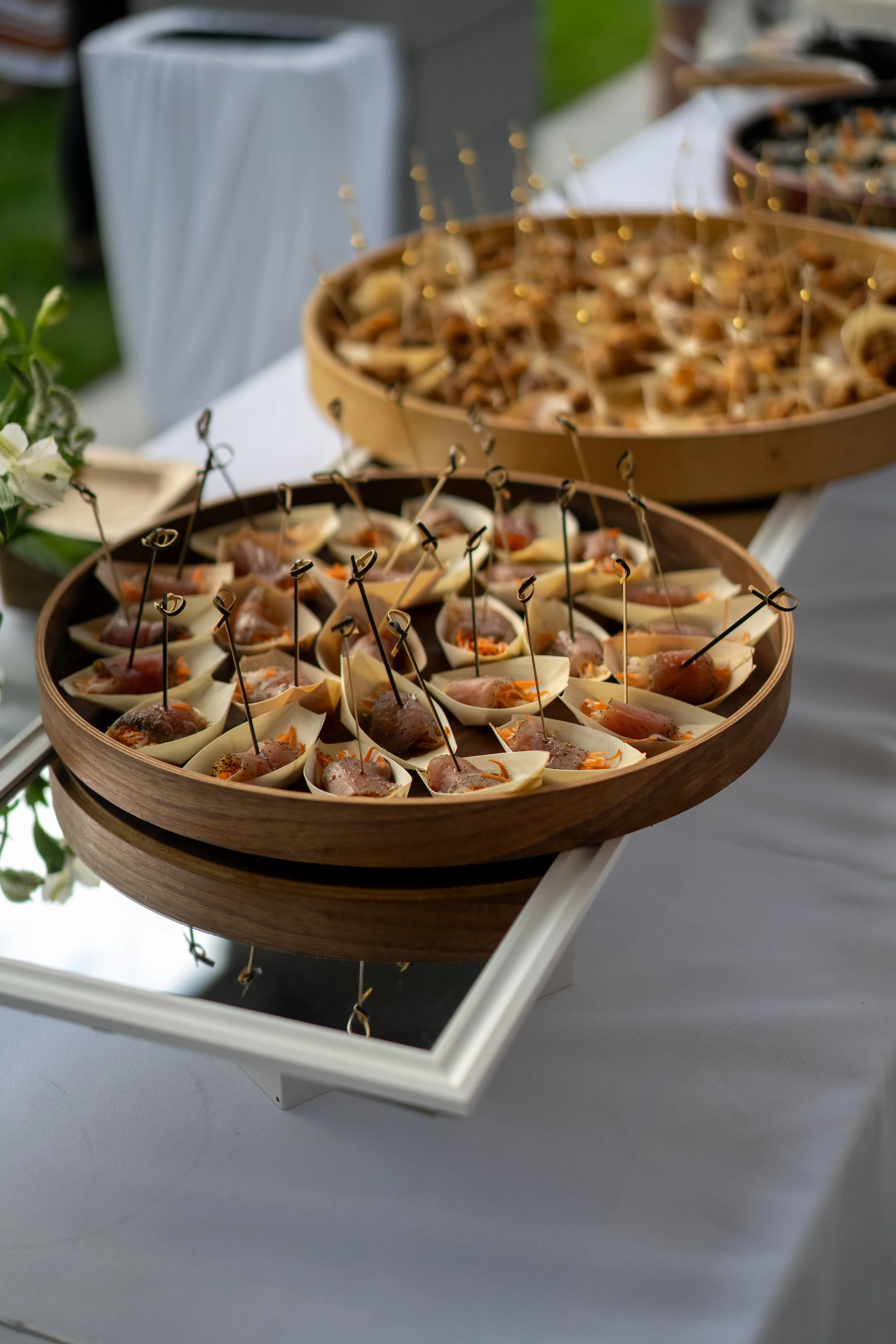 Two wooden trays filled with small appetizers arranged on a white stand at an outdoor event.