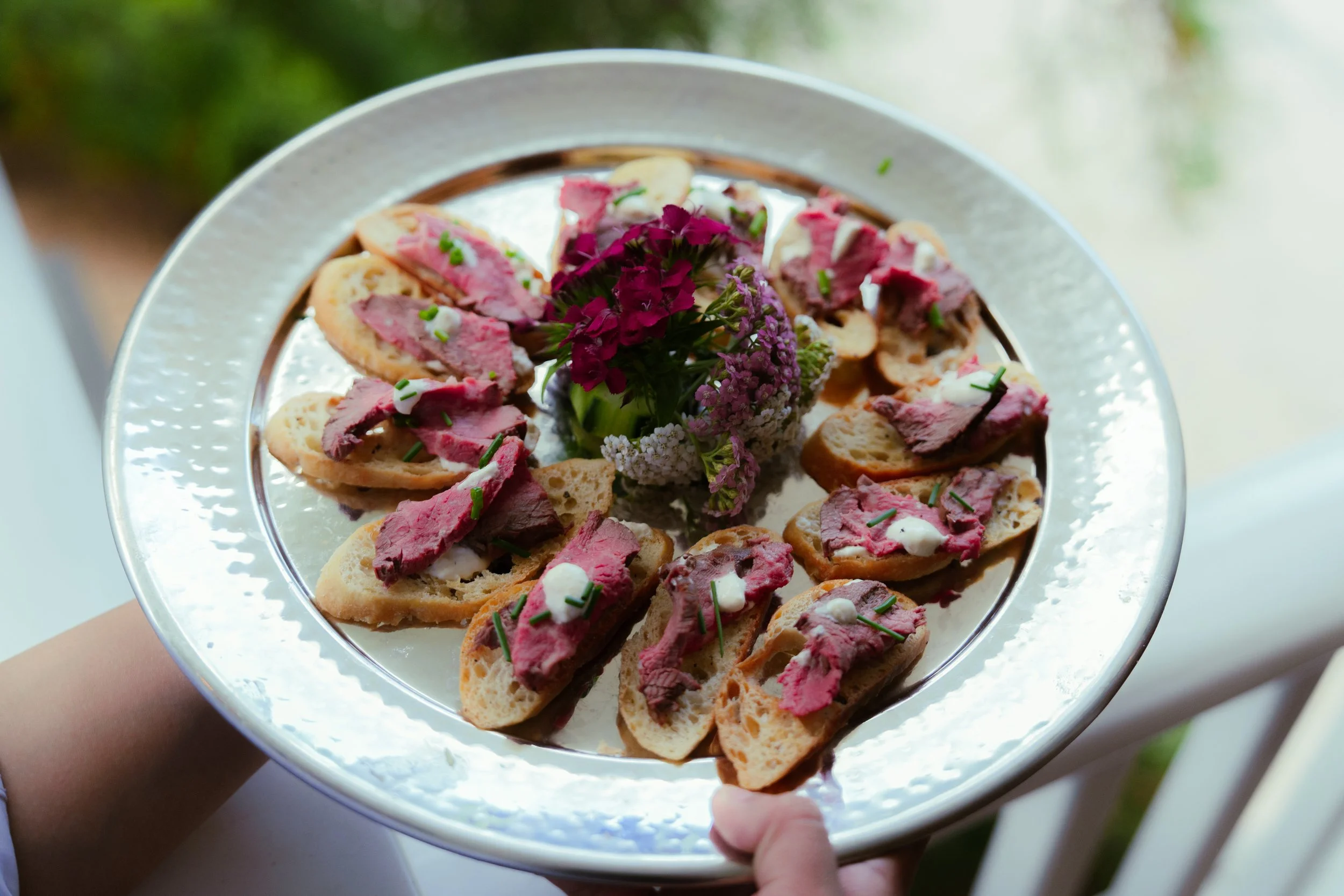 A silver platter with slices of bread topped with sliced roast beef, garnished with chopped chives and small dollops of creamy sauce, centered with a small bunch of purple flowers.