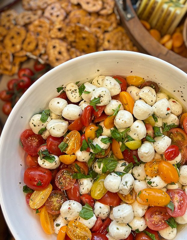 Caprese salad with cherry tomatoes, fresh mozzarella balls, basil, and seasoning in a white bowl, with cookies and snacks in the background.