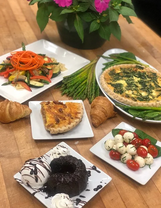 Assorted dishes on a wooden table including a vegetable salad, grilled quiche, tomato and mozzarella salad, fresh asparagus, spinach tart, and two scoops of ice cream with chocolate sauce.